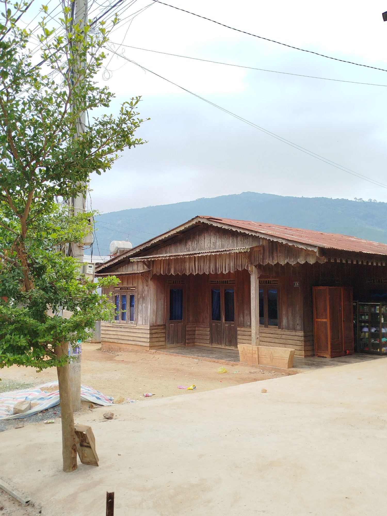 A single-story wooden house with a rusty corrugated metal roof sits in front of a low mountain under a cloudy sky in Đarahoa, Vietnam. The house has a porch with wooden supports and multiple windows with blue curtains visible. A wooden cabinet stands against one wall. A green tree is on the left side of the image, partially obscuring a utility pole with numerous wires attached. The ground in front of the house is dirt and concrete.