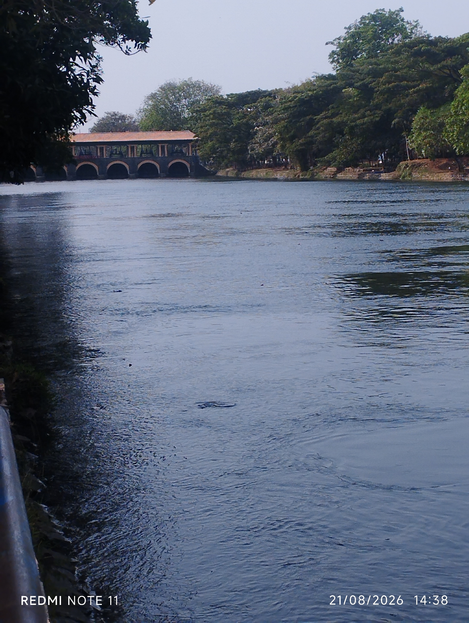 The image depicts a body of water, likely a river or lake, with a building structure featuring arched openings in the background. Dense green foliage lines the opposite bank, contributing to a naturalistic setting. The water's surface exhibits ripples, suggesting a gentle current or wind. In the foreground, a dark railing or barrier is partially visible on the left side. Text overlays indicate "REDMI NOTE 11" and a date and time stamp of "21/08/2026 14:38". The location is stated to be Karawang, Indonesia, however, there is no specific visual information within the image to confirm this geographical context.