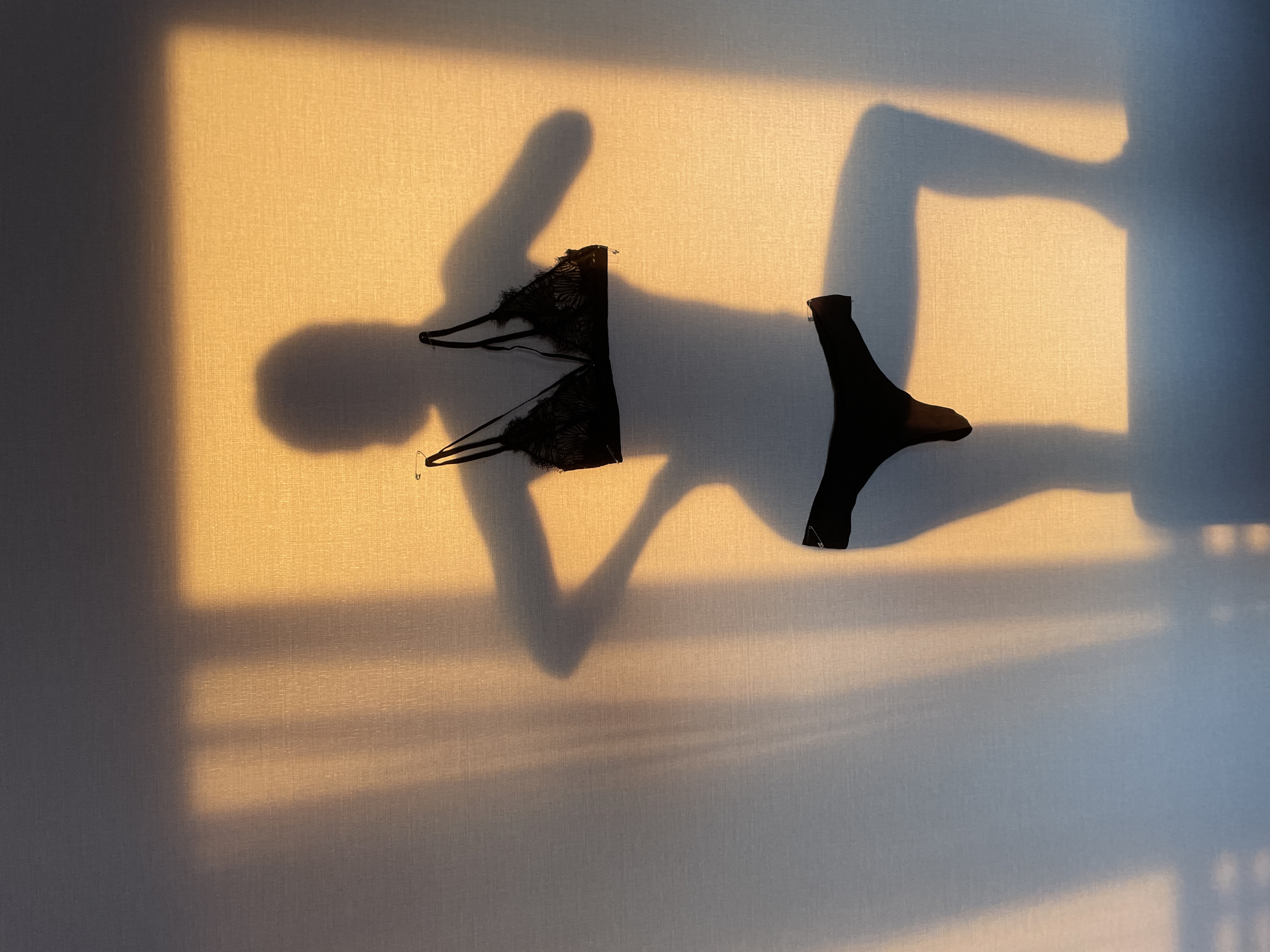 The image shows a beige wall with shadows and two articles of black lingerie. A window's shadow is visible in the upper left and lower right corners. The lingerie consists of a bra on the left, pinned to the wall, and a thong on the right, also pinned. A person's shadow is cast on the wall in the center, appearing to be a woman in a provocative pose. The location in Подшивалово, Russia, is not discernible from the image.