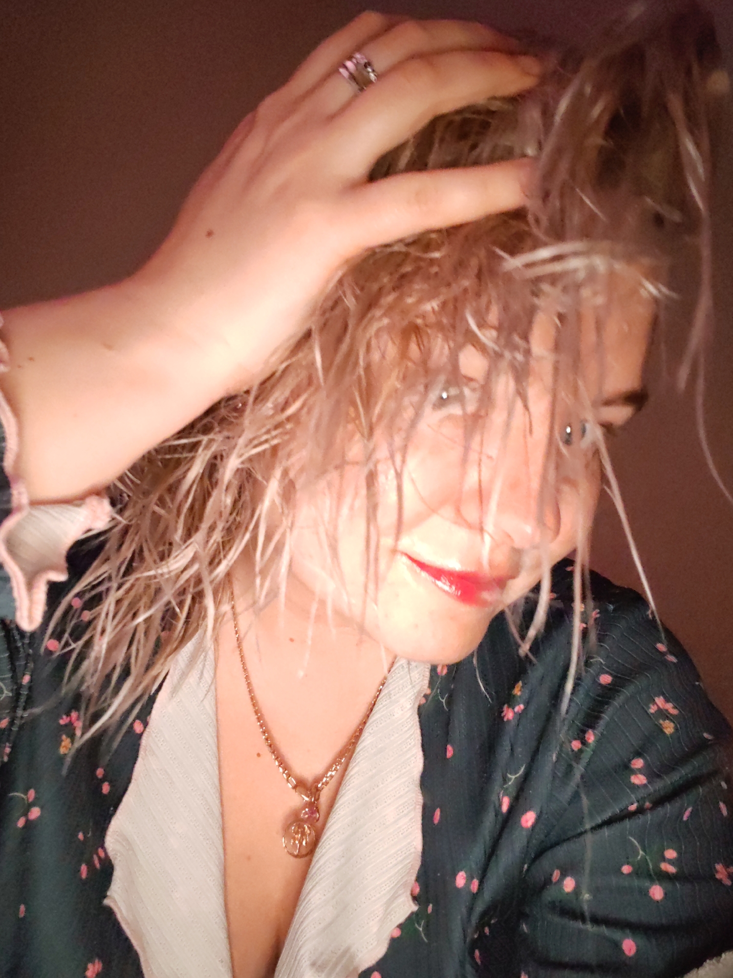 A close-up shot shows a woman with damp, light-colored hair partially obscuring her face. She is wearing red lipstick and has a hand on her head, with a ring visible on one finger. She is wearing a dark-colored robe or top with a floral pattern over a light-colored undershirt or garment. A gold chain necklace with a pendant is visible around her neck. The background appears to be a dark, indistinct color. There is no indication of a location in Izhevsk, Russia.