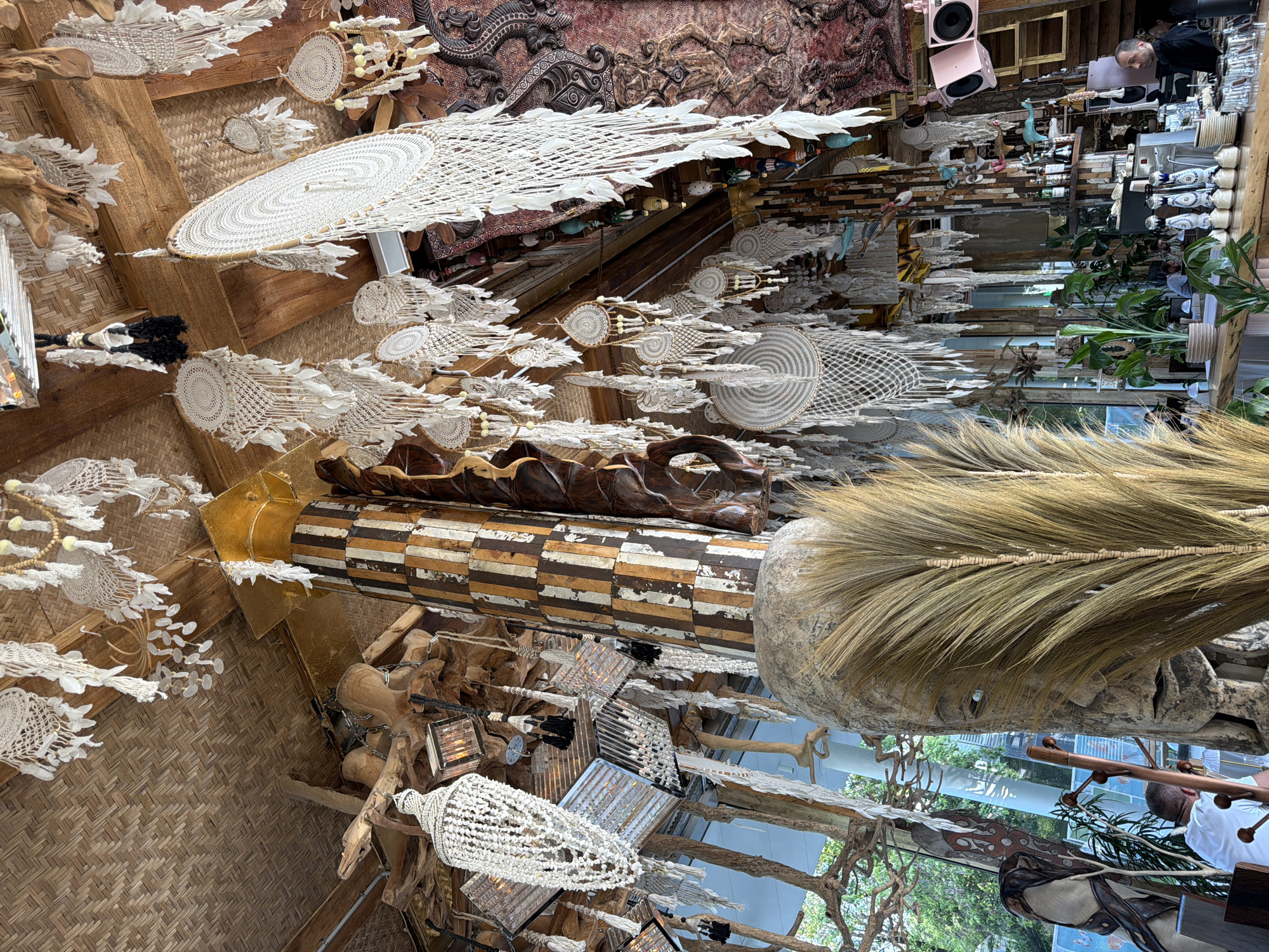 The image captures the intricately decorated interior of an establishment in Moscow, Russia, possibly a restaurant, bar, or shop. The scene is dominated by a bohemian and rustic aesthetic, characterized by a wealth of natural materials and handcrafted elements.
Numerous white and off-white macrame-style decorations are suspended densely from the ceiling, which is composed of wooden beams and a woven bamboo or wicker texture. These hangings feature a variety of circular patterns, intricate netting, and delicate fringes, some adorned with small white, leaf-like shapes or beads, and a few with yellow or black accents.
Prominent architectural and decorative features include large, carved wooden elements. A distinctive cylindrical pillar is wrapped in distressed, striped wood panels of brown, white, and black. Other organic, gnarled wooden pieces, resembling branches or roots, are interspersed throughout. A large, rounded object covered in dried straw or grass also adds to the natural texture. On an upper wall, an elaborate, dark-toned carved wooden panel or tapestry with complex patterns and figures contributes to the rich, textured environment.
In the upper right, a man with short dark hair and a beard, wearing a dark top, stands behind a counter, likely a bartender or staff member. He is looking towards the left. Behind him, shelves are stocked with bottles, glasses, and ceramic items, while two pale pink speakers are mounted higher up. Another person in a white t-shirt is partially visible further down.
The space is lit by a combination of natural light, likely filtering through unseen windows and reflecting off glass surfaces, and artificial light from decorative chandeliers made of small white beads or shells. Green potted plants introduce touches of foliage. The overall impression is one of an artfully curated and textured space, exuding a warm and inviting atmosphere. No discernible text is visible within the image.