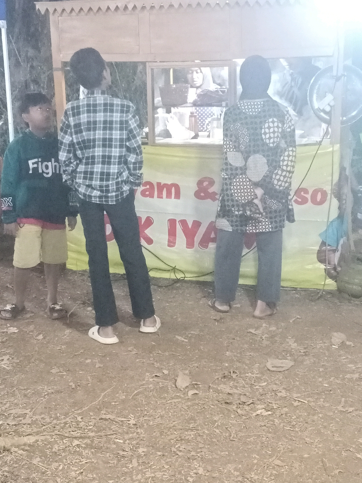 The image captures an outdoor scene in Tasikhargo, Indonesia, likely at a food stall or market. In the foreground, two young boys stand on the dirt ground. The boy on the left wears a dark green hooded sweatshirt with white text reading "Fight" and yellow shorts. He is looking up and to his left. The boy on the right wears a blue and white plaid long-sleeved shirt and dark pants.
Behind the boys, a food stall structure is visible. A yellow banner with red lettering is stretched across the front of the stall. Partially visible text on the banner includes "&" and "SO". In the background, a person wearing a dark head covering and a patterned tunic stands facing away from the camera, looking towards the stall. Another person, wearing a headscarf, is partially visible behind a glass display inside the stall. The environment appears to be a public area with loose dirt and scattered leaves on the ground, suggesting a somewhat rustic or informal setting. The lighting suggests it may be evening or dusk.