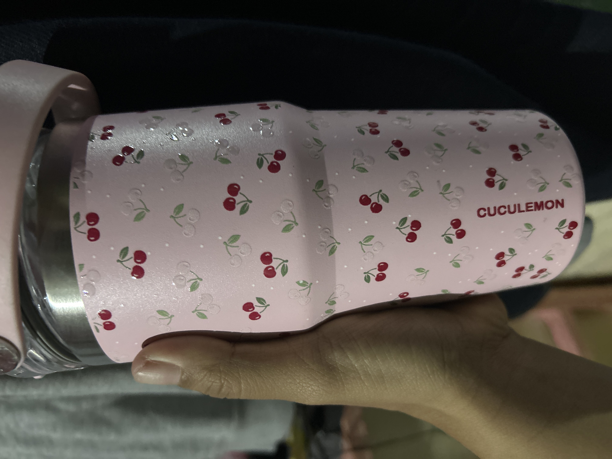 A hand, with visible creases on the palm and fingers, holds a pink insulated tumbler adorned with a pattern of cherries and small white dots. The tumbler has a metallic rim and a pink lid with a handle, suggesting it is a travel mug. The background is dark and out of focus, with hints of gray fabric on the left. The lighting is somewhat dim. The text "CUCULEMON" is visible vertically on the tumbler.