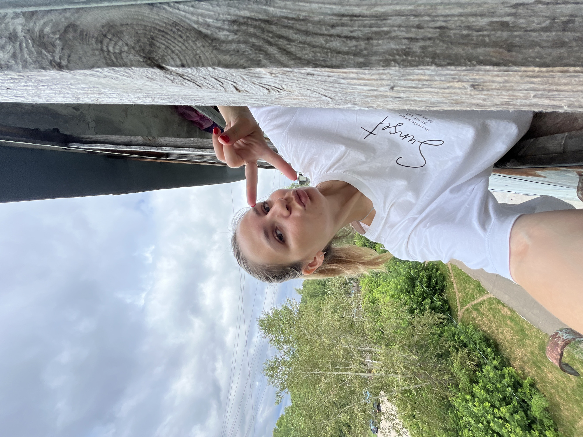 A young woman is taking a selfie from an elevated position, leaning over a wooden surface. She is wearing a white t-shirt with the word "Sunset" printed on it in cursive. She has blonde hair pulled back in a ponytail and is making a peace sign with her right hand, her nails painted red. Her lips are pursed, and she has a playful expression. In the background, there are green trees under a cloudy sky, with power lines visible. The scene suggests it is daytime, possibly late afternoon given the "Sunset" on her shirt. The image is oriented upside down in relation to the landscape.