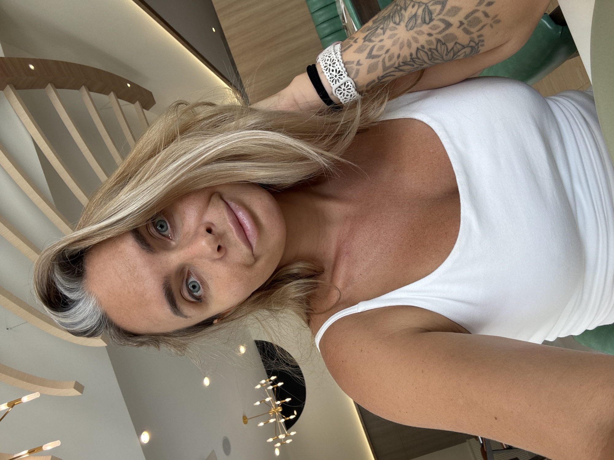 A woman with blonde hair and blue eyes is taking a selfie in what appears to be a restaurant. She is wearing a white tank top. She has a tattoo on her right arm. In the background, there is a modern light fixture and wooden architectural elements, suggesting an indoor dining establishment. The location provided, Khok Kham Subdistrict, Thailand, is an industrial area known for its factories and shipping ports, which does not align with the visual cues of a restaurant environment.