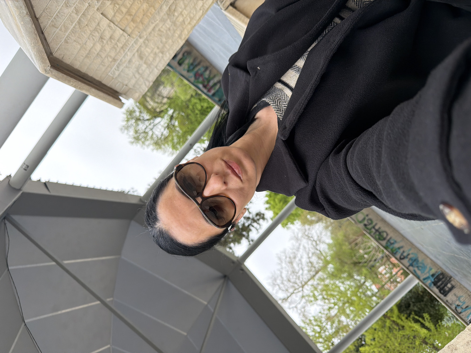 This media file captures a rotated, close-up selfie of a woman outdoors in Pleven, Bulgaria. The woman, positioned in the lower left of the frame, has dark hair and is wearing dark sunglasses with brown frames, obscuring her eyes. Her lips are slightly parted, revealing a hint of color. She is dressed in a dark, textured coat or jacket, possibly black, layered over a lighter, checkered or patterned garment. A decorative brown button is visible on her right sleeve cuff.
The background is an outdoor setting under an overcast, light grey sky. To the upper left, there is a light-colored, textured stone or concrete structure, possibly part of a building or monument. Below this, modern grey architectural elements, characterized by angular panels, stretch across the mid-ground, suggesting a canopy or shelter. Lush green foliage, including trees with some bare branches and bushes, indicates a spring or early summer season. A prominent grey concrete beam or pillar diagonally traverses the mid-right background, covered in stylized graffiti text. Due to the image's rotation and the graffiti's nature, the text is mirrored and largely illegible, though some letter-like shapes such as "PHEJ" and other indistinct markings can be seen. The overall scene suggests an urban or park environment.