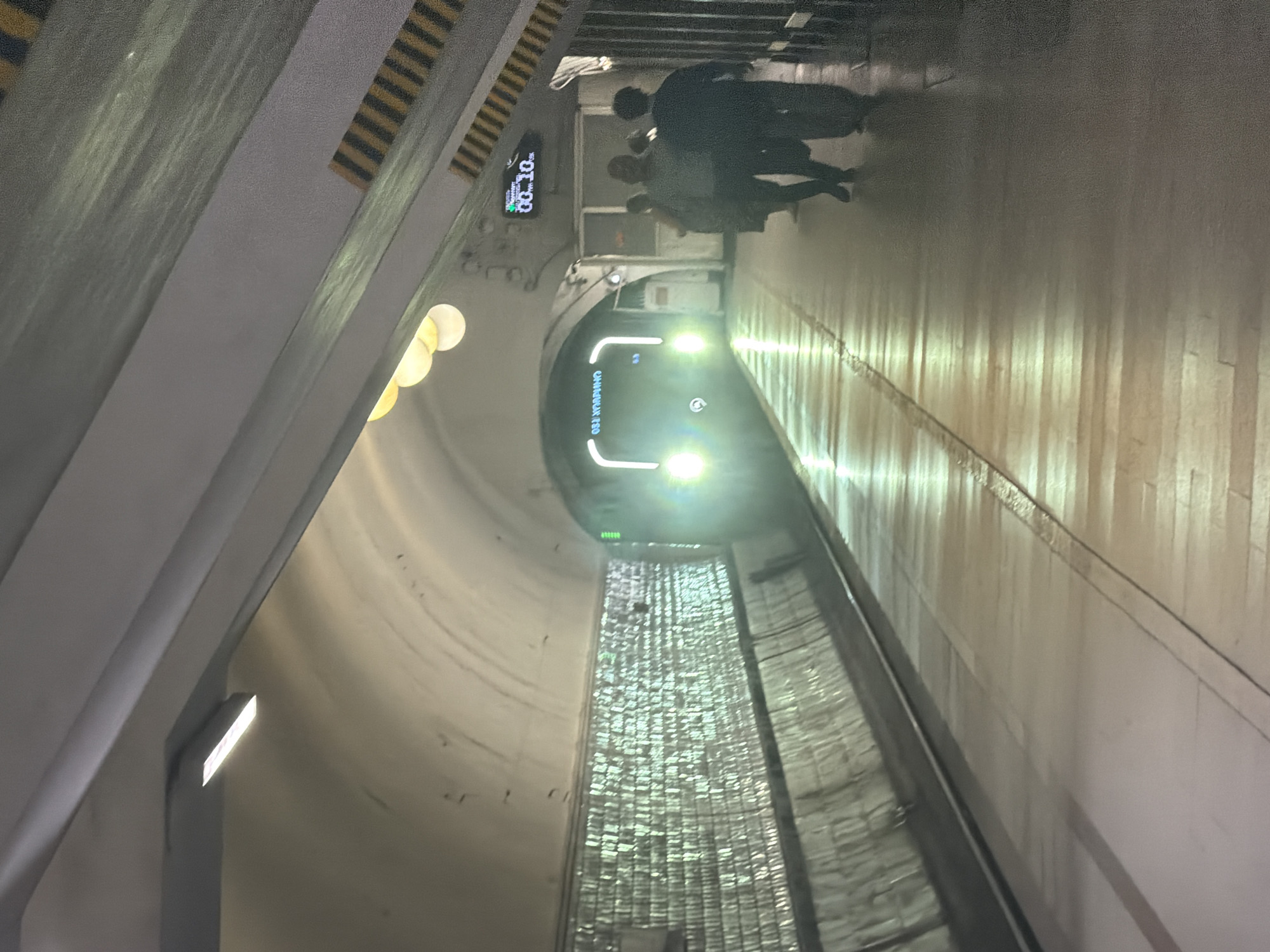 The image depicts a subway station with a train approaching. The station has a curved ceiling with overhead lighting. The walls on the left are covered in reflective, textured material. The platform is paved with wooden planks on the left and a darker material on the right.
In the distance, a train with its headlights on is entering the station from a tunnel. The train has the word "OESL HAXPHIO" written on its front in white, illuminated lettering. A digital display on the wall above the platform shows "00.10".
On the right side of the platform, three individuals are walking away from the camera. The person in the foreground is wearing a dark top and pants, the person in the middle is wearing a grey top and dark pants, and the person behind them is wearing a dark top and dark pants. They are all facing away from the approaching train. The overall lighting is dim, with the train's headlights and the platform lights being the primary sources of illumination.