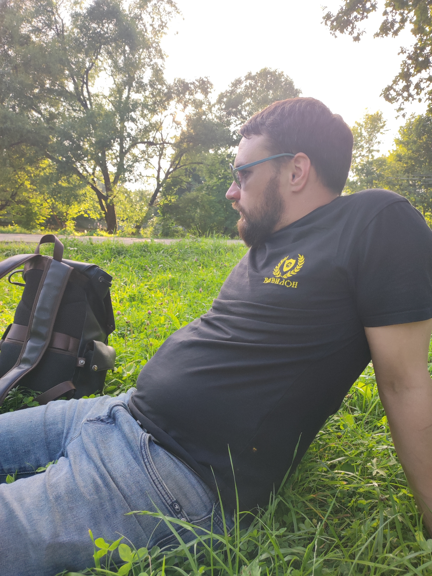 A medium shot captures a man sitting in a grassy area, facing right and looking away from the camera. He is wearing a black t-shirt with a yellow emblem and text that appears to read "BABVLON," and light blue jeans. A black backpack with brown accents rests behind him. The background is a sun-dappled park with trees and a paved path. The lighting suggests it is daytime, possibly late afternoon, given the warm glow. The overall scene depicts a casual outdoor moment in a park. The location is identified as Alabino, Russia.