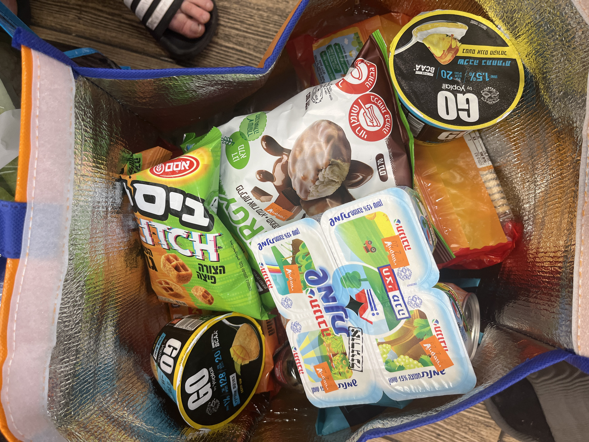 The image shows the contents of a shopping bag filled with various food products, mostly Israeli brands.
The brands that can be identified are:
* Osem Bissli snacks
* Yoplait GO yogurt cups
* Tnuva dairy products
* Chocolate-covered rice crackers.
The bag appears to be an insulated cooler bag. Part of someone's foot wearing an Adidas sandal can be seen in the upper left corner of the image. The location is likely Tel Aviv, Israel, based on the Hebrew text and brands visible on the food items.