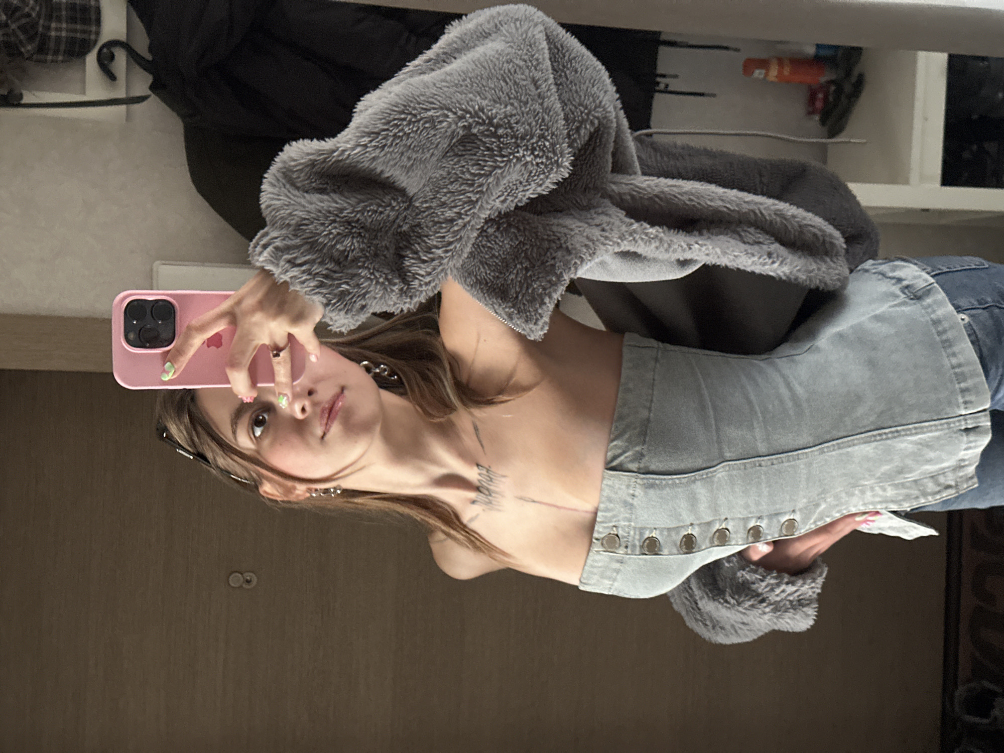 A young woman is taking a mirror selfie in what appears to be a domestic environment. She is standing, wearing a light blue denim corset top with four visible buttons and a grey fuzzy jacket with large sleeves. Her hair is brown and appears to be tied back. She is holding a pink smartphone in her right hand, which is angled to capture her reflection. The background shows a brown door with a small circular fixture, a white shelf with some items on it, and a black garment hanging on a hook. The overall lighting suggests an indoor setting, possibly a room in an apartment in Moscow, Russia.