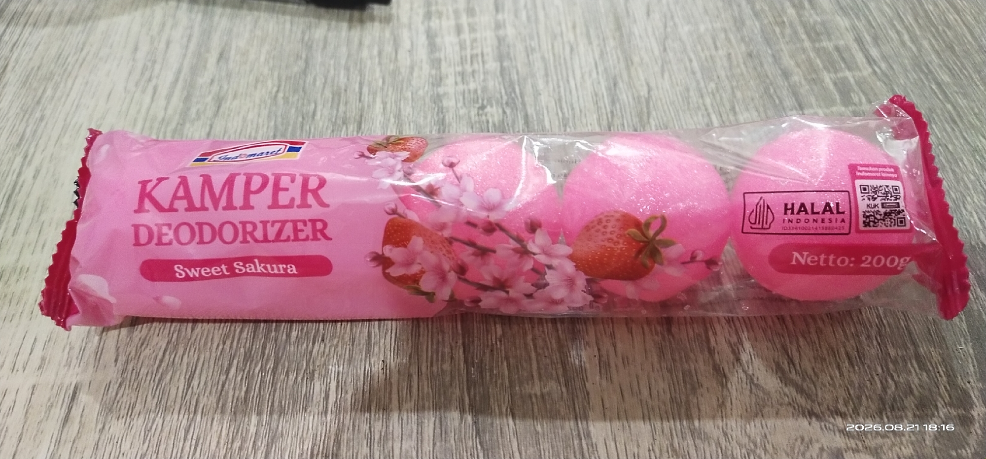 The image shows a package of three pink, egg-shaped air fresheners with a "Sweet Sakura" scent. The packaging is predominantly pink and features images of strawberries and cherry blossoms. Text on the package indicates it is "KAMPER DEODORIZER" and has a net weight of 200g. A "HALAL INDONESIA" certification and a QR code are also visible. The air fresheners are resting on a wooden surface. The timestamp in the lower right corner of the image indicates the photo was taken on 2026.08.21 at 18:16. There is no discernible information or context relating to the city of Matabu, Indonesia, within the provided image.
