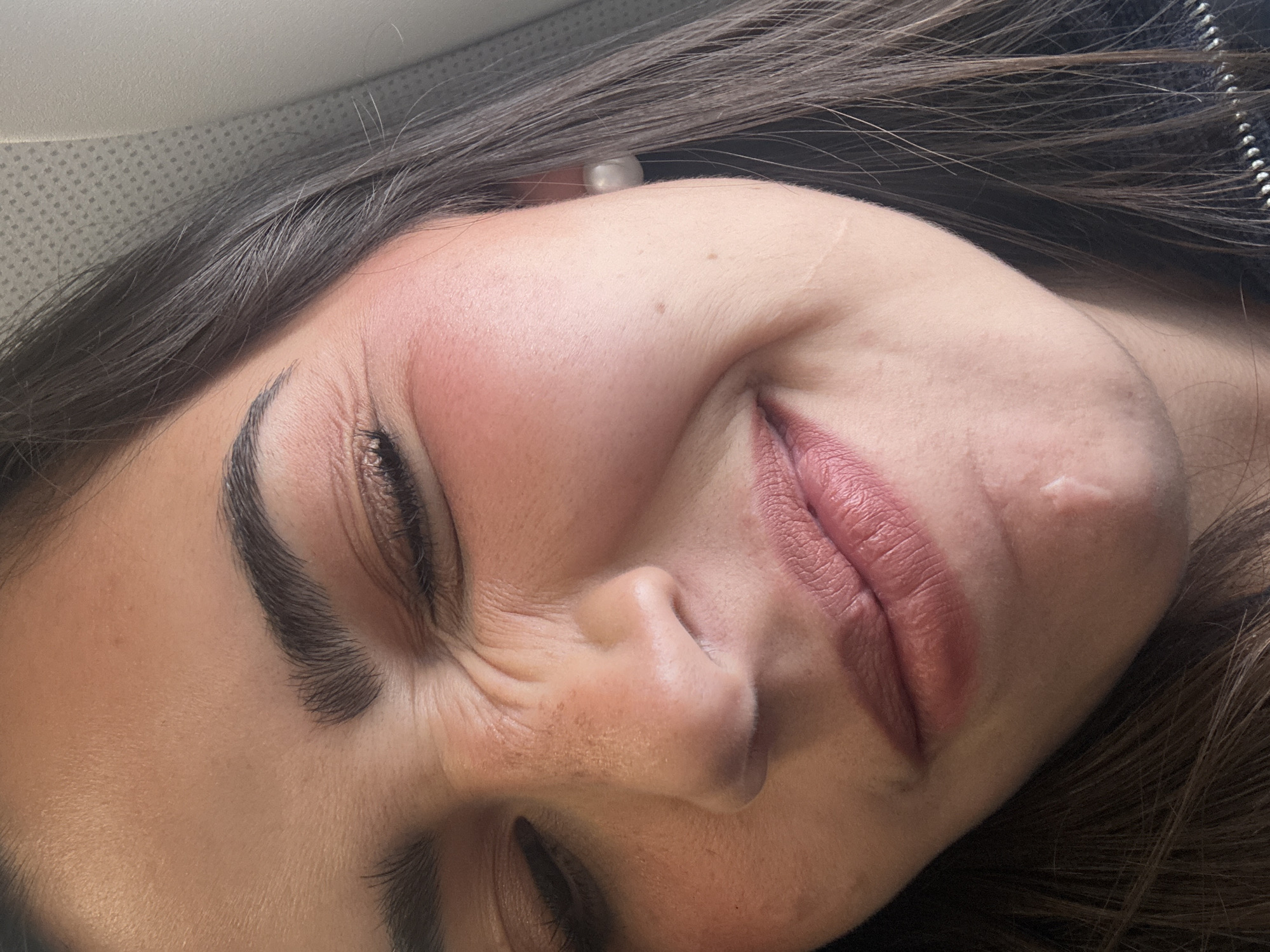 A close-up, angled view of a woman's face, likely taken indoors. The woman has dark hair and fair skin, with noticeable makeup, including defined eyebrows, blush on her cheeks, and lipstick. Her eyes are mostly closed, and her face is scrunched up, suggesting she is squinting or smiling widely. There is a small blemish or scar visible on her right cheek. The background is blurred, but appears to be a light-colored surface. There is no information about the location, or what city or country it was taken in.