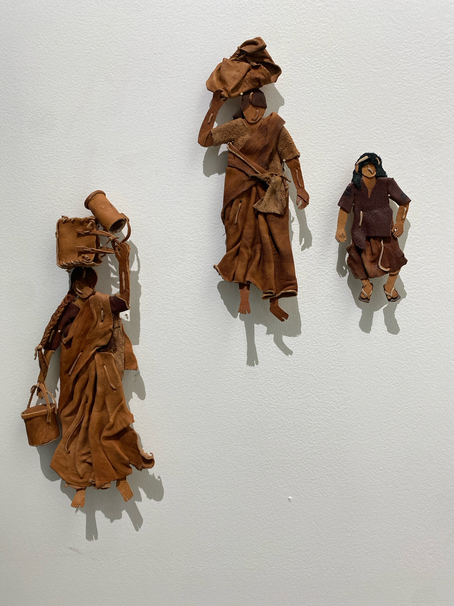 This is a photograph of three leather art figures mounted on a textured white wall. The figures appear to represent people, possibly women, in traditional attire.
The figure on the left is the largest and is shown in profile, facing right. It is carrying a cylindrical object and a square bag on its head. It also carries a small bucket in its left hand. The figure's body is draped in a brown leather garment, and its legs and feet are exposed. Its right arm is raised, supporting the objects on its head, and its left arm hangs down. A braided pigtail extends from the back of its head.
The figure in the middle is positioned higher and slightly to the right of the first figure. It is also shown in profile, facing right. This figure is carrying a bundle on its head. It wears a brown leather garment that drapes over its body, and its arms and legs are visible.
The figure on the far right is the smallest and is positioned slightly higher than the middle figure, to its right. It is shown facing forward, with its arms at its sides and its legs exposed. This figure wears a dark brown tunic and a skirt, and appears to have sandals on its feet. It has dark hair.
The figures are all made of what appears to be distressed or aged leather, giving them a rustic and textured appearance. They are mounted on the wall at varying heights and angles, casting distinct shadows below them. The wall appears to be clean and evenly lit, with some texture visible. The overall impression is of an art installation.
There is no information provided about the location in the city of Khimki, Russia, within the image or its description.