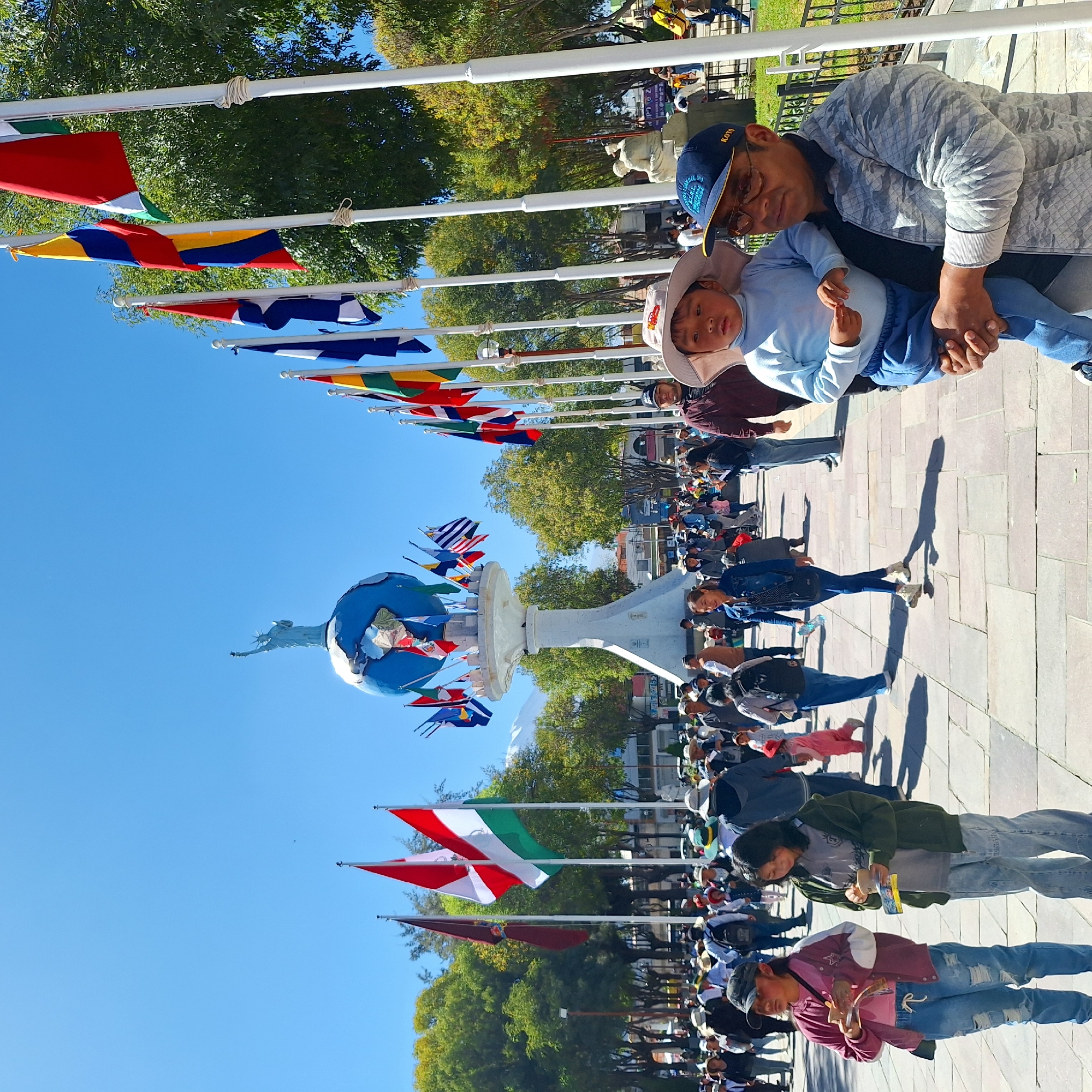 The image shows a crowded outdoor scene, likely a public plaza in Cerro Colorado, Peru, based on the presence of a monument topped with a globe and the flags of various nations displayed along the plaza. Many people, dressed in casual clothing, are walking and interacting. A man is holding a child, who is wearing a hat. The weather appears to be sunny and clear. The monument is surrounded by trees and buildings.