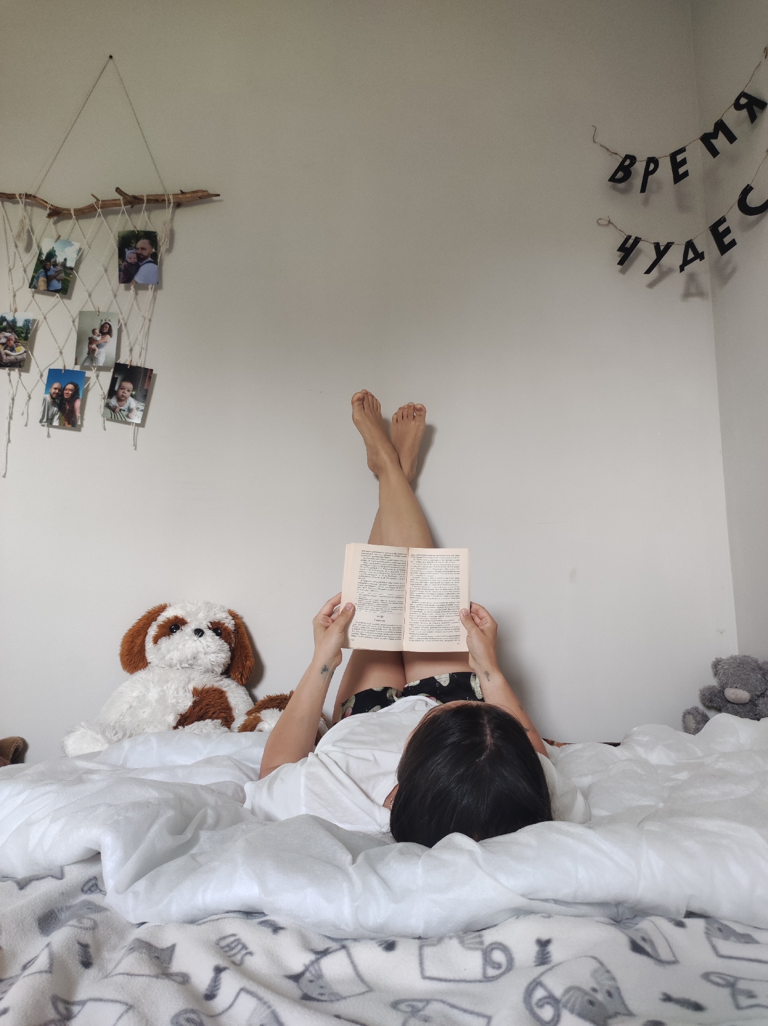 A person is lying in bed, reading a book with their legs propped up against a white wall. To the left of the person is a stuffed dog, and to the right is a gray teddy bear. Above the dog is a homemade photo display hanging from a branch, and above the bear is a banner in a language that is not English. The bed is covered in a white comforter with a gray blanket at the foot of the bed. The room is located in the city of Unknown, Unknown.