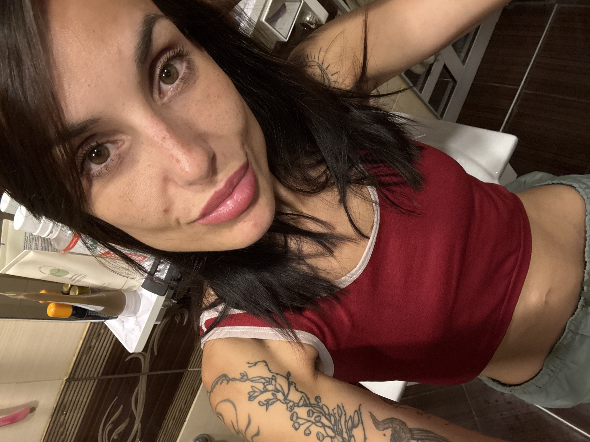 A young woman with dark hair and brown eyes is taking a selfie in what appears to be a bathroom. She is wearing a red tank top and grey sweatpants. There are tattoos on her left arm and shoulder. In the background, a shelf holds various toiletries, including tubes of lotion and a toothbrush. The walls are tiled with a brown and beige pattern.