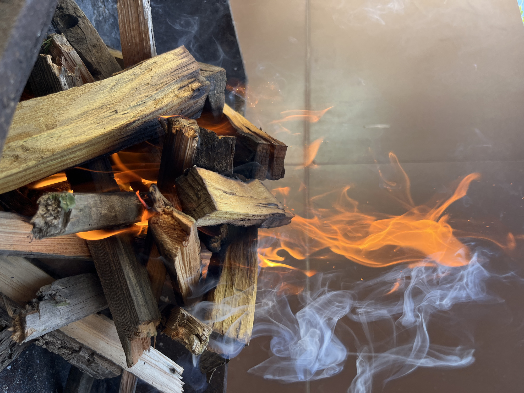 A pile of wood is stacked and partially on fire. Flames and smoke rise into the air. The location is likely a controlled fire pit. The background is a dark, reflective surface. The scene appears to be indoors. It does not appear to be in the city of Kaspiysk, Russia.