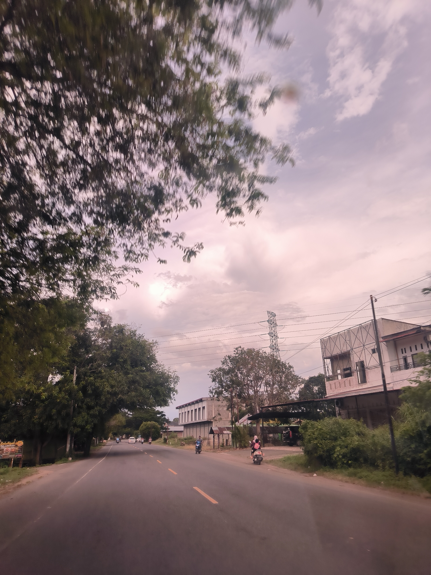 This outdoor image captures a road scene, likely taken from a moving vehicle, given the motion blur visible in the foreground trees and sky. The scene is set in Reuhattuha, Indonesia.
The primary subject is a paved road stretching into the distance, with faint yellow double lines marking the center and single yellow lines on the sides. Lush green trees line both sides of the road, particularly dense on the left, with their upper branches heavily blurred across the top and upper left of the frame. The sky is overcast with a mix of light and darker grey clouds, suggesting either an approaching storm, early morning, or late afternoon light.
On the right side of the road, several buildings are visible, some appearing residential and others possibly commercial or under construction with exposed frameworks. A tall metal lattice transmission tower and numerous power lines traverse the sky above these buildings. Further down the road, traffic includes several motorcycles; one with two riders (a person in a pink top and another in a dark top, both wearing helmets) is visible closer on the right, and other motorcycles and a white car can be seen in the distance.
On the left side of the road, partially obscured by trees, there's a sign with visible Indonesian text. The words "Selamat" and "PELAMINAN" are discernible, suggesting it might be a welcome sign related to a wedding or event, as "pelaminan" refers to a wedding dais. The general atmosphere is that of a typical, active road in a semi-urban or rural setting.