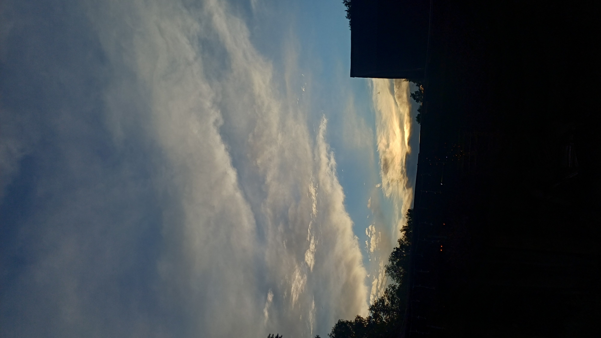 The image depicts a sky with predominantly cloudy conditions. The clouds range from shades of white to gray, with some areas illuminated by a golden light, possibly from the setting sun. The sky is blue in sections where clouds are less dense. In the lower portion of the frame, there is a dark, silhouetted area indicating either trees or buildings, which obscures the view of the landscape. The image is rotated counter-clockwise. Based on the provided context, it may be inferred that this scene is located in or near Timokhovo, Russia, but no specific location details are discernible from the image itself. There are no people or identifiable subjects visible. The environment is characterized by the sky and the dark, silhouetted elements in the foreground.