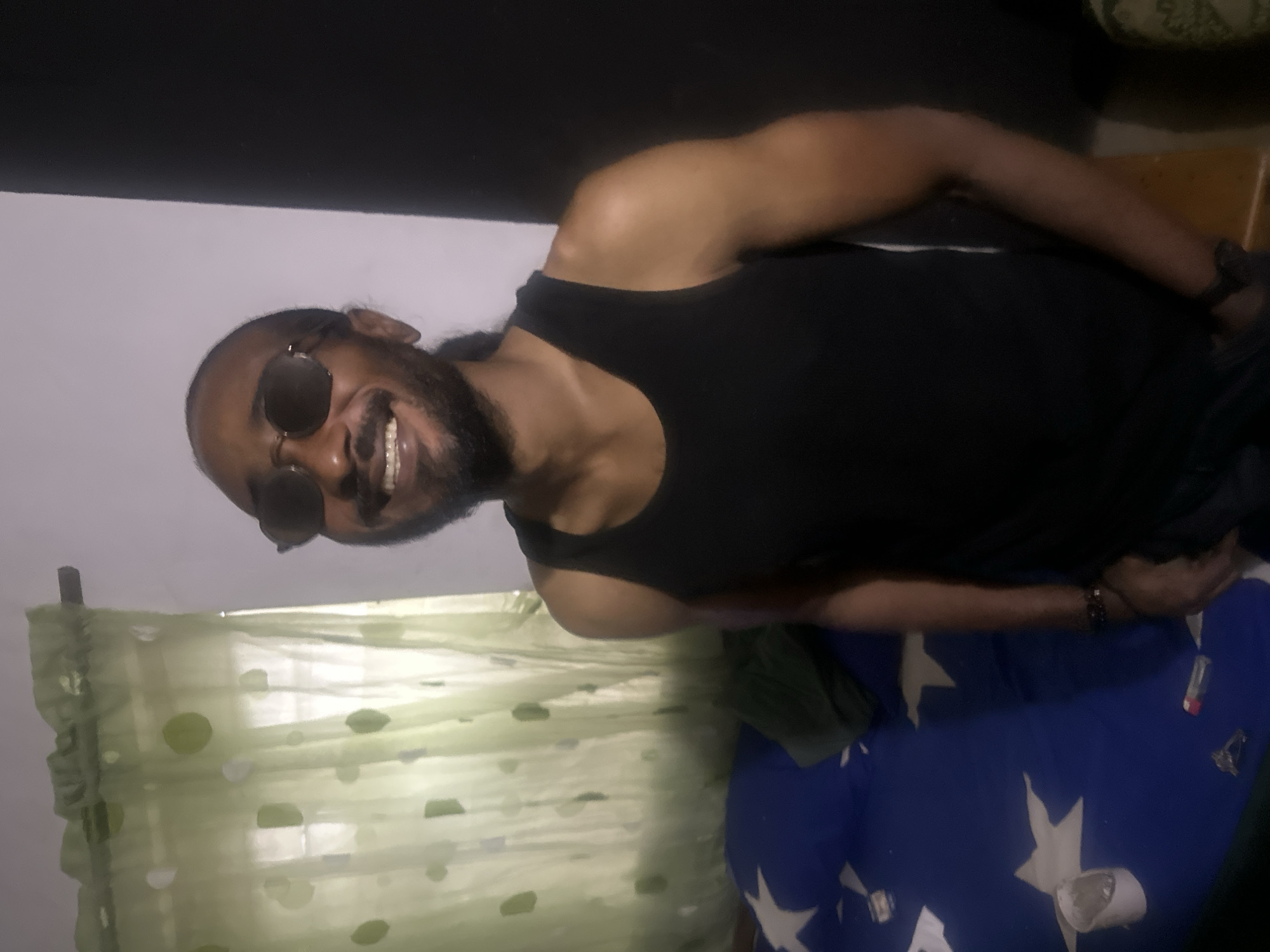 The image captures a man in what appears to be an indoor setting in Jos, Nigeria. He is positioned horizontally, looking directly at the camera with a wide, joyful smile that reveals his teeth. The man has a dark skin tone, a beard, and long dark hair, possibly tied back. He is wearing dark sunglasses that cover his eyes and a plain black tank top. A bracelet is visible on his right wrist.
To the man's left (which would be below him if the image were upright), a sheer, light green or white curtain with various circular patterns (some solid, some outlined) is visible. Below the man (to his right in an upright view), there is a blue fabric, possibly a blanket or sheet, decorated with large white star patterns. Several small, indistinct objects, including what resembles a lighter, are resting on this blue fabric. The background features a dark, possibly black, surface and a lighter-colored wall. The lighting suggests an indoor environment, making the time of day unclear. The man's expression conveys happiness and amusement. No discernible text is visible in the image.
