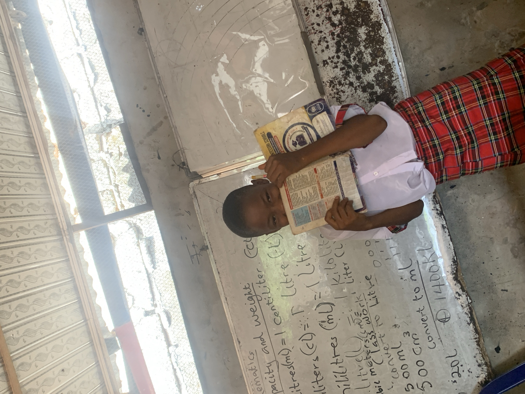 A young person in a white shirt and red plaid skirt is holding books in front of them, obscuring their face. The person is standing next to a white chalkboard with handwriting on it. The handwriting discusses mathematics, capacity, and weight, including conversions of liters to milliliters and other units. The words "MONTESSORI SCHOOLS" are visible on the book. This image was captured indoors, in what looks like a classroom setting. It's not possible to determine the exact location in the city of Ukwu-Abwa, Nigeria from the image alone.