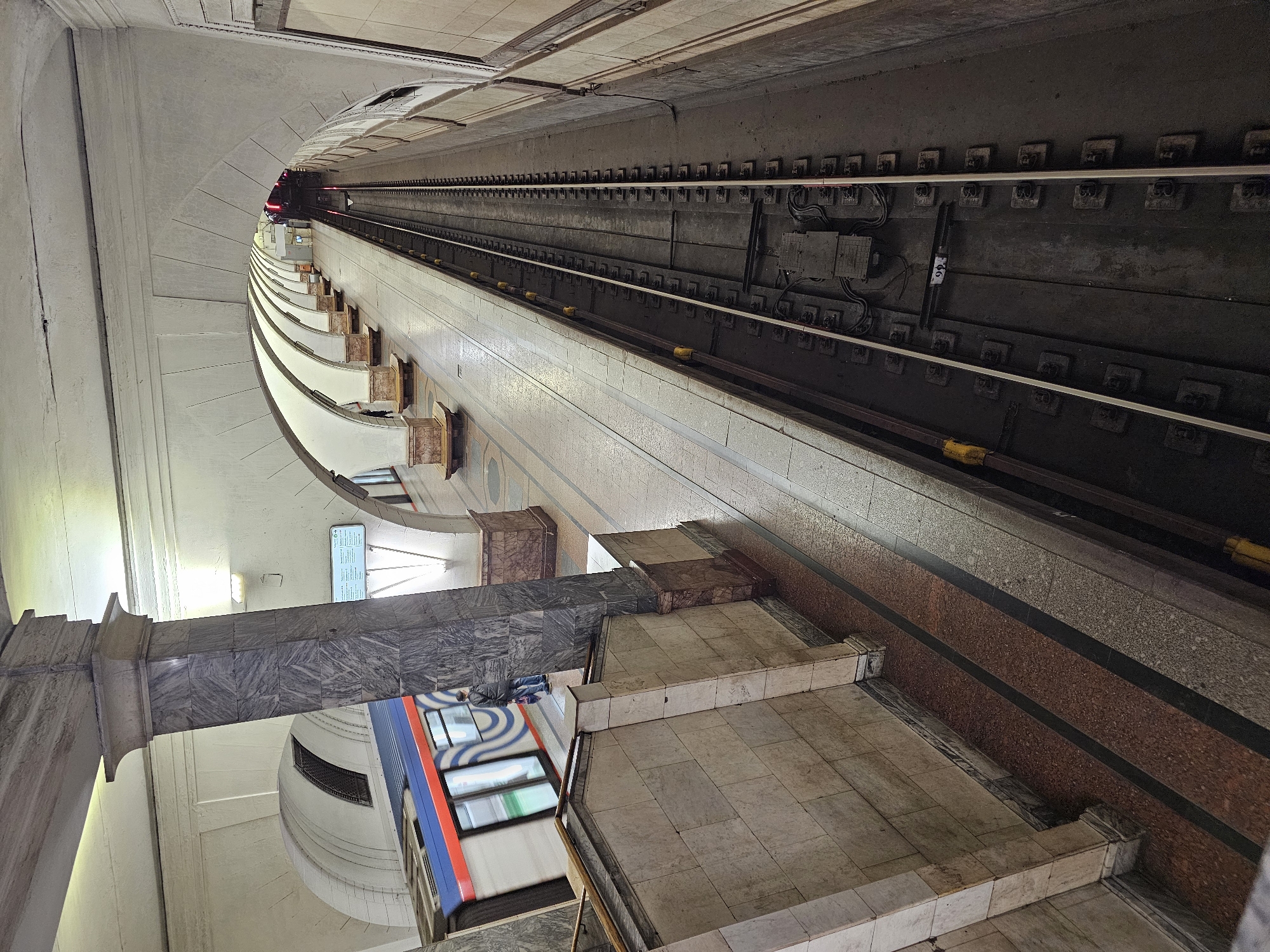 The image shows a subway station in Moscow, Russia. The perspective is angled, looking down the platform and tracks. A train with a blue and red exterior is partially visible. The station architecture includes marble columns, tiled walls, and curved archways. The tracks and related infrastructure are visible, leading into the distance.