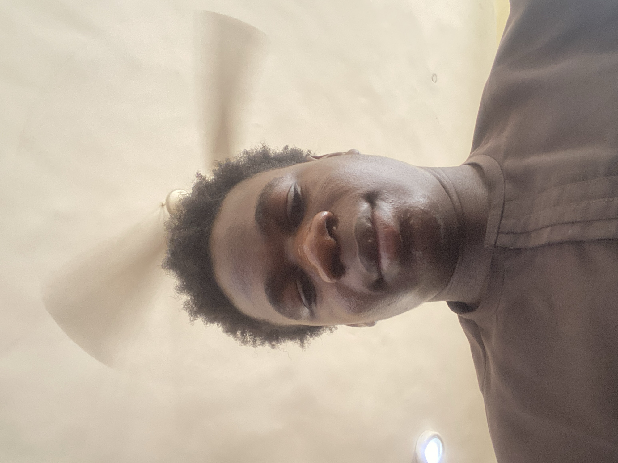 A low-angle shot depicting a Black male indoors. He has short, curly black hair and a brown shirt. Above him is a white ceiling fan and a ceiling light. The location is unspecified, but given the prompt, it can be inferred to be Mararaba, Nigeria.