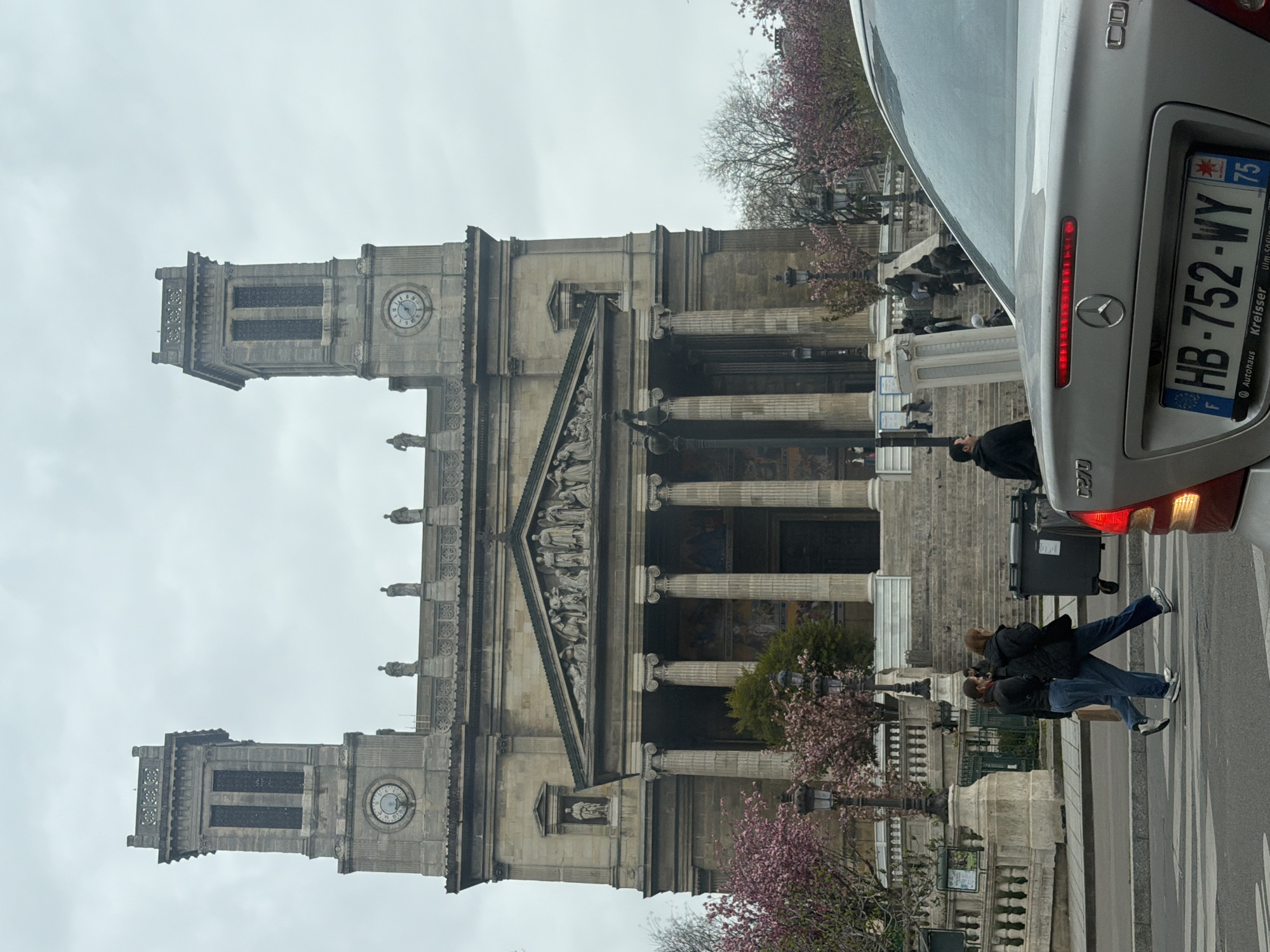 The image shows the Saint-Vincent-de-Paul Church in Paris, France. The church has a classical facade with columns and two towers. A silver Mercedes-Benz is visible on the right side of the frame with a license plate reading "HB-752-WY 75." The sky is overcast. Several people are visible: Two individuals are walking on the sidewalk in front of the church, and another person is standing near a trash bin on the steps. Cherry blossom trees are in bloom. The scene is taken from a street-level perspective.