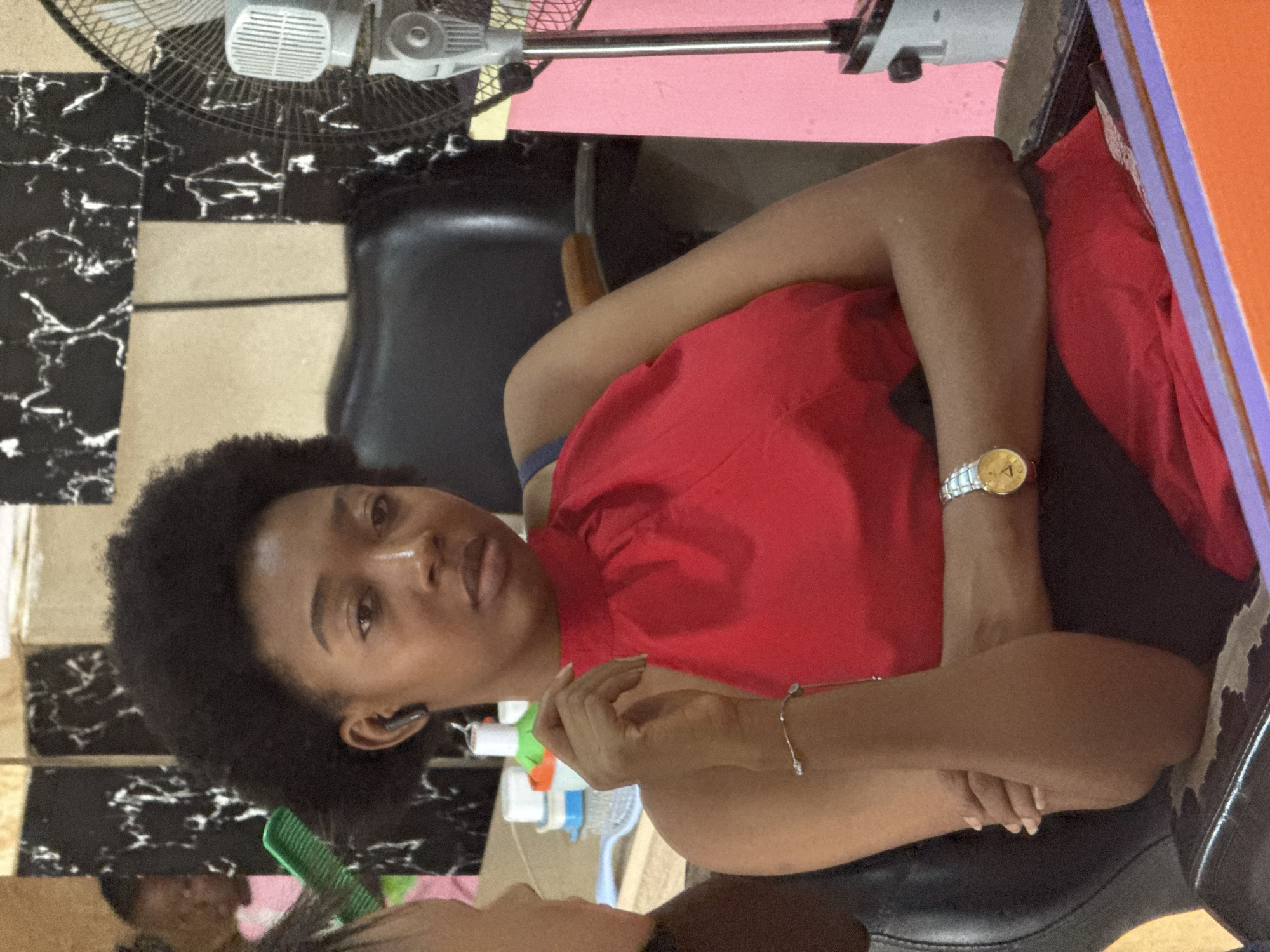 A woman with short, dark curly hair sits in a salon, wearing a red gown over black clothing, with a gold watch and silver bracelet on her left arm. She is seated in a black chair with a textured seat, and is looking directly at the camera. Her arms are crossed in her lap. In the foreground, another person is visible near a green comb and salon bottles. In the background, a black and white patterned wall is visible near a white electric fan. The location is inferred to be in Abuja, Nigeria.
