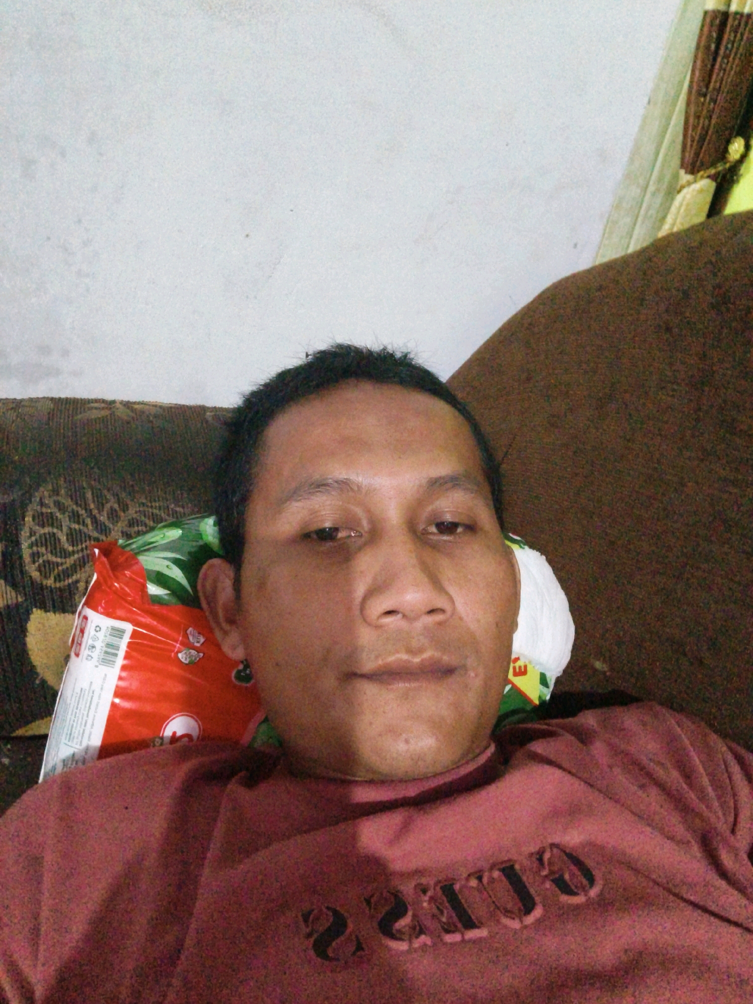 A man is lying on a couch, looking at the camera. He has short black hair and brown eyes. He is wearing a maroon shirt with the word "GUESS" printed on it. Next to him is a red and green bag. The wall behind him is white. The couch is brown and has a patterned fabric. The lighting is dim. The image appears to be taken indoors. The image does not contain any identifiable landmarks or specific details that would allow for pinpointing a location within Banyuwangi, Indonesia.