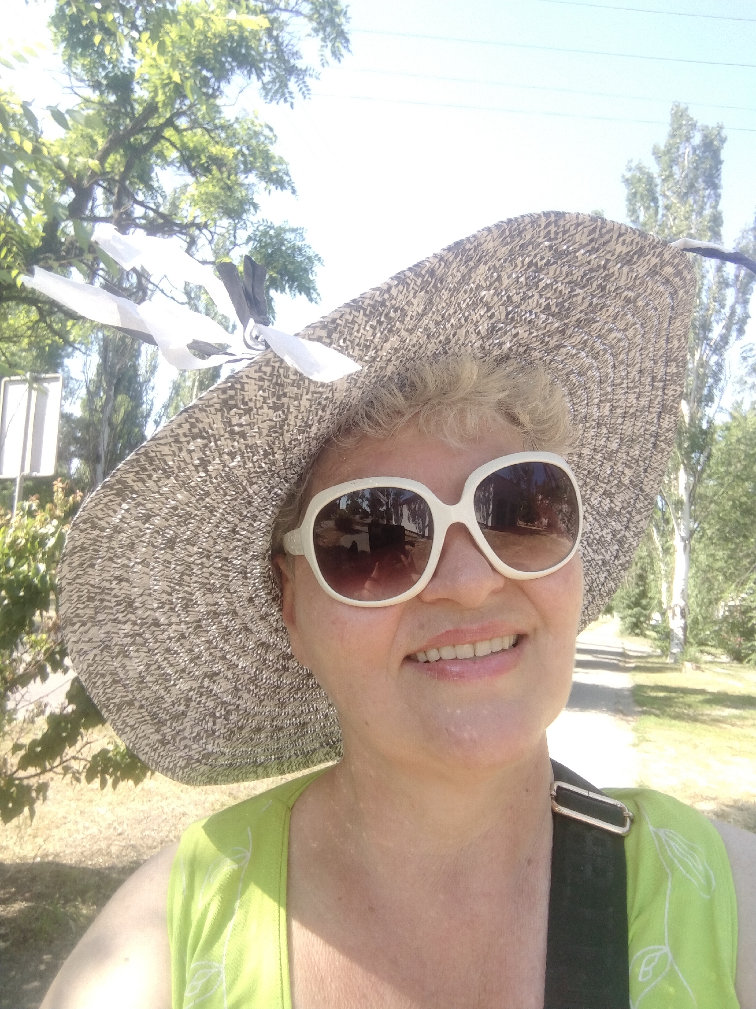 A woman is taking a selfie outdoors in Horishni Plavni, Ukraine, on a sunny day. She is wearing a wide-brimmed straw hat with a black and white ribbon, large white-rimmed sunglasses, and a bright green top with a white floral pattern. She has blonde hair and is smiling. The background shows trees, a blue sky, and a path.