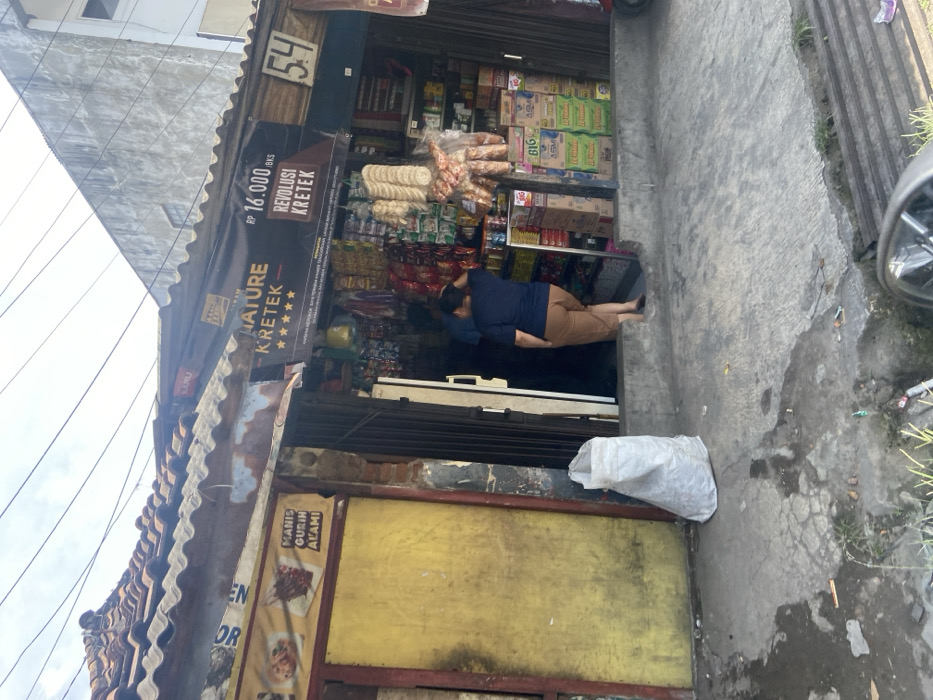 The image shows the front of a small shop in Kecamatan Dayeuhkolot, Indonesia, at latitude -6.966 and longitude 107.592. The shop is located on a street corner and has a yellow wall with a sign that reads "Manis Gurih Alami." The shop is open and a woman in a blue shirt and brown pants is walking out of the shop. There are many products on the shelves inside the shop, including snacks, drinks, and other household goods. There is a white bag on the ground outside the shop. The shop is in a residential area with a cloudy sky above.