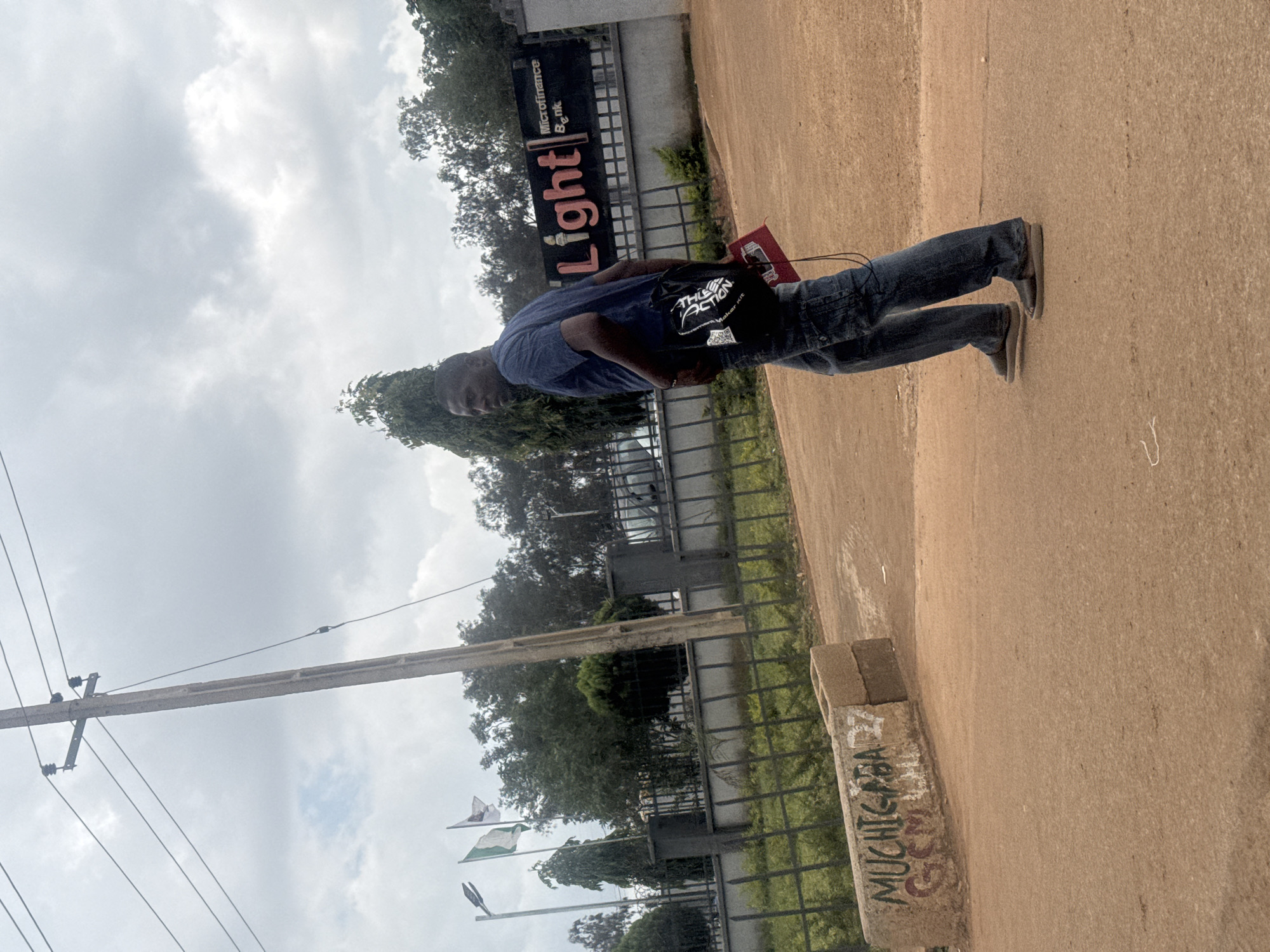 A man is standing outside, leaning against a wall. He is wearing a blue t-shirt, blue jeans, and grey shoes. He has a black bag slung over his shoulder and is holding a red book in his left hand. The man is looking towards the camera.
In the background, there is a sign that reads "Microfinance Bank" and "Light." There are also trees and a fence. The sky is cloudy, suggesting it might be an overcast day. The ground is made of a reddish-brown dirt. There are flags visible in the distance.
The overall scene appears to be in an outdoor urban or semi-urban environment. The graffiti on the concrete pillar reads "MUCHIGBA".