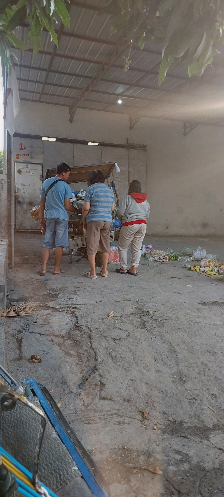 In the city of Semarang, Indonesia, an outdoor market scene is depicted. Three individuals are present. On the left, a man wearing a blue t-shirt and denim shorts stands facing a food cart. To his right, a woman in a striped blue and white t-shirt and brown shorts is positioned behind the food cart. Further to the right, another person in a grey hoodie with a red lining and light-colored pants stands behind the cart. The food cart appears to be laden with various goods, including what look like onions or garlic. The ground is paved with concrete, showing some wear and tear. Scattered around the area are plastic bags, some containing produce, and a broom lies on the ground. The scene is set under a corrugated metal roof supported by exposed beams, suggesting an open-air market or a covered stall. A white door with the number "1A" and the word "BERCKEL" is visible on the left side of the image. The lighting suggests it is daytime, with sunlight filtering in, creating some lens flare.