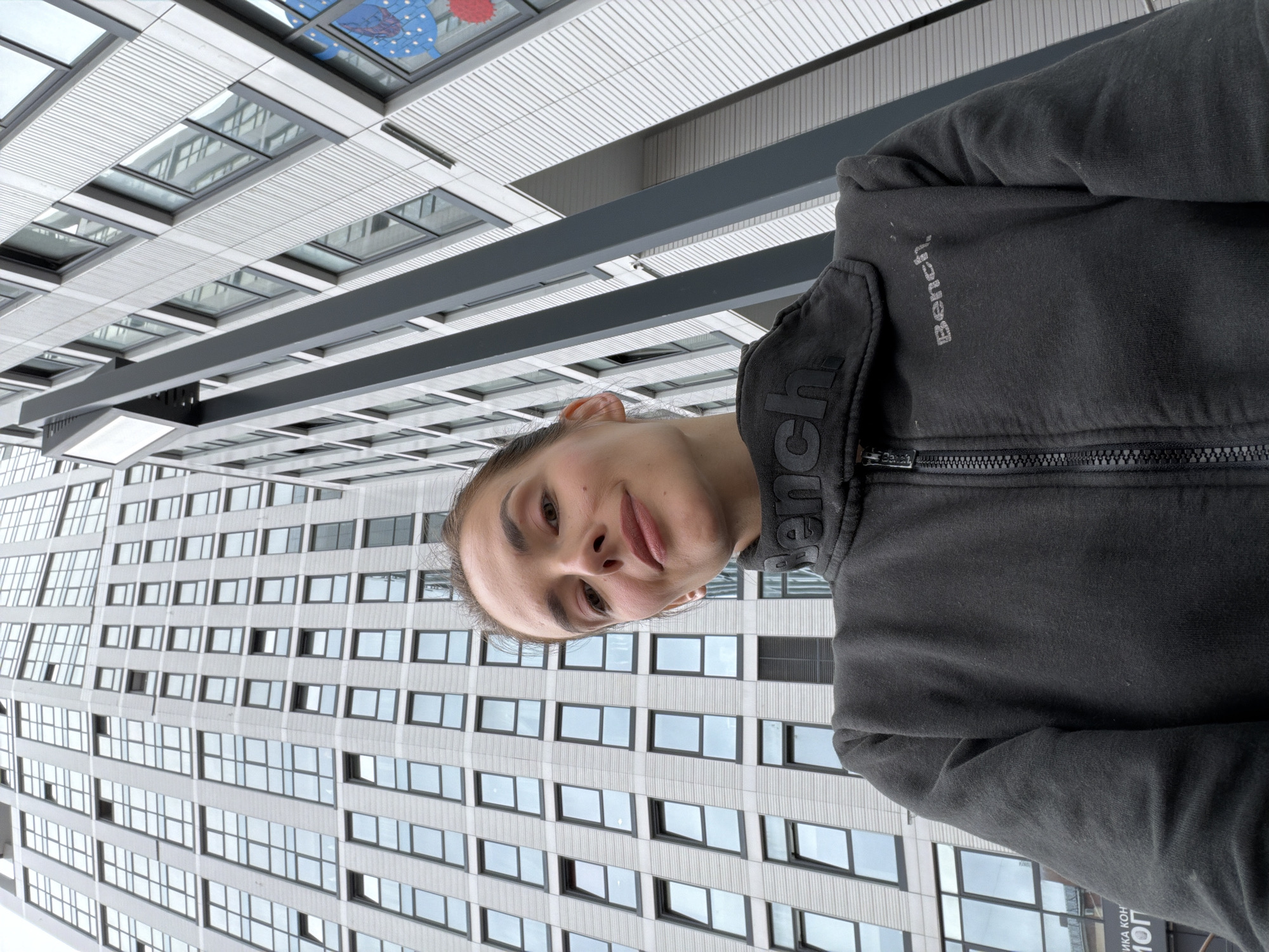 A woman is taking a selfie in front of a modern high-rise building in Ufa, Russia. The building is tall and has many windows, and its architecture is angular and contemporary. The woman is wearing a dark grey zippered jacket with the brand name "Bench" visible on the chest. She is looking towards the camera with a slight smile. The perspective of the photo is looking up at the building, making it appear even larger. The sky is overcast, contributing to the cool-toned lighting of the scene. The woman's hair is pulled back, and she has visible makeup on her face.