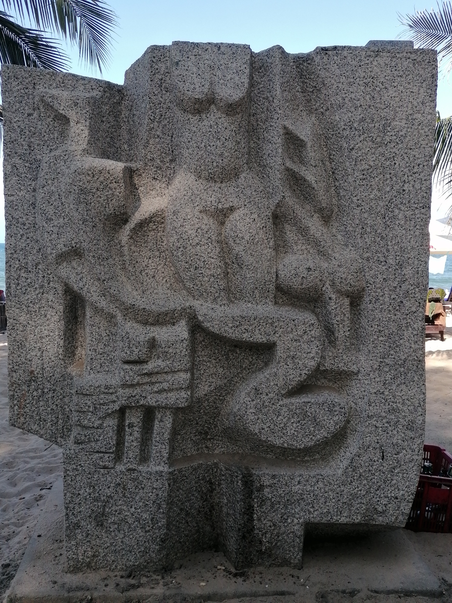 The image shows a large, grey granite sculpture on a sandy beach. The sculpture features abstract, carved shapes, including what appears to be a figure with rounded elements and linear features. The beach is located in Nha Trang Ward, Vietnam. The sea and sky are visible in the background, along with palm trees. A red plastic crate sits near the base of the sculpture. Other beach furniture can be seen in the background.