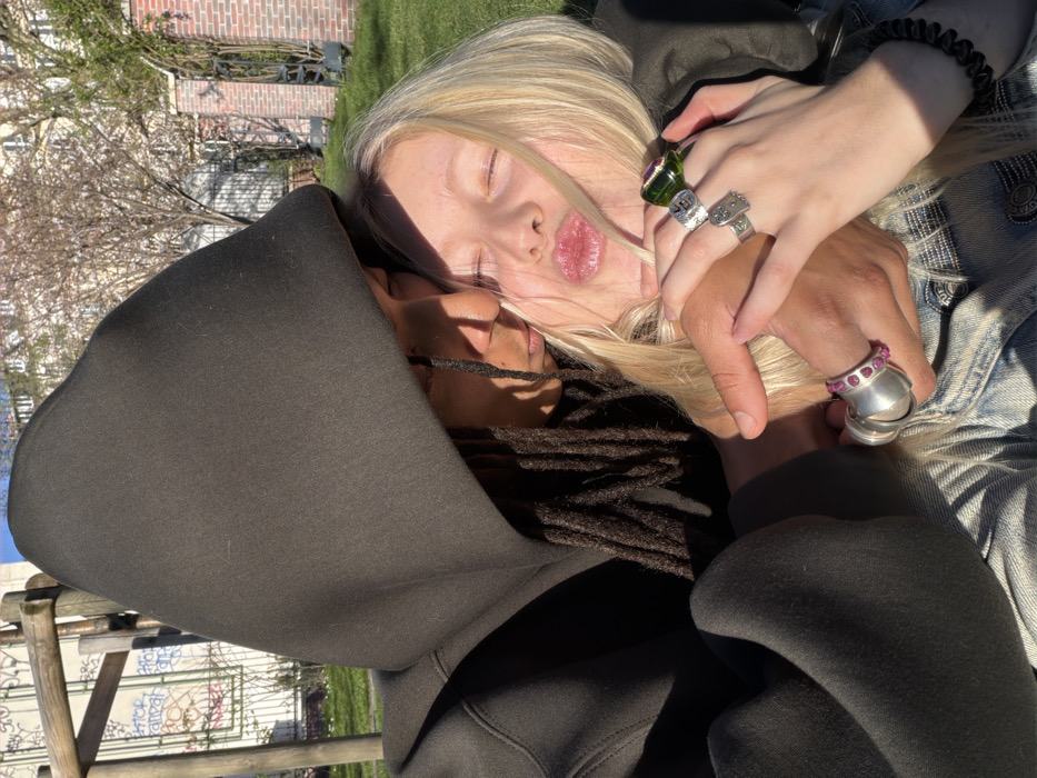 This outdoor, eye-level image captures two individuals in a park environment, consistent with Parisian urban green spaces. One individual, with dark skin and dreadlocks, wears a dark hooded sweatshirt with the hood up, embracing a blonde individual. The hooded subject's face is positioned near the blonde subject's head. The blonde individual has closed eyes and puckered lips, and is wearing a light blue denim jacket, partially visible. Their hands are interlocked, displaying multiple rings: the darker-skinned individual wears a large silver ring with red stones on their left ring finger and a large smooth silver ring on their left middle finger. The blonde individual wears a substantial green stone ring on their right ring finger, and two silver rings with engraved patterns on their right middle and index fingers. A black spiral hair tie is visible on the blonde subject's right wrist. The background features a grassy area, a wooden park structure to the left, and a light-colored building with graffiti on its lower facade. To the right, a brick wall with decorative ironwork is present, characteristic of Parisian city gardens. Several bare-branched trees are visible under direct sunlight.