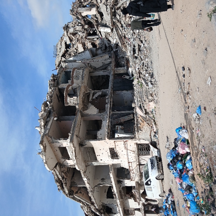 The image depicts a scene of severe urban destruction in Jabalia, Palestinian Territory, under a partially cloudy blue sky. A multi-story concrete building, heavily damaged and partially collapsed, stands in the left-center of the frame. Its upper levels are largely destroyed, revealing fractured concrete and exposed rebar. The exterior walls display numerous small circular perforations consistent with bullet impacts. To the right and behind this structure, a substantial pile of building rubble extends. On the unpaved ground in the left foreground, multiple large accumulations of colorful plastic trash bags are visible, along with scattered refuse. A white commercial van is parked adjacent to the base of the damaged building on the left. Two individuals are observed walking away from the camera on a dirt path: one male wearing dark clothing and carrying an object in his right hand, and one female in a long black garment and head covering.
