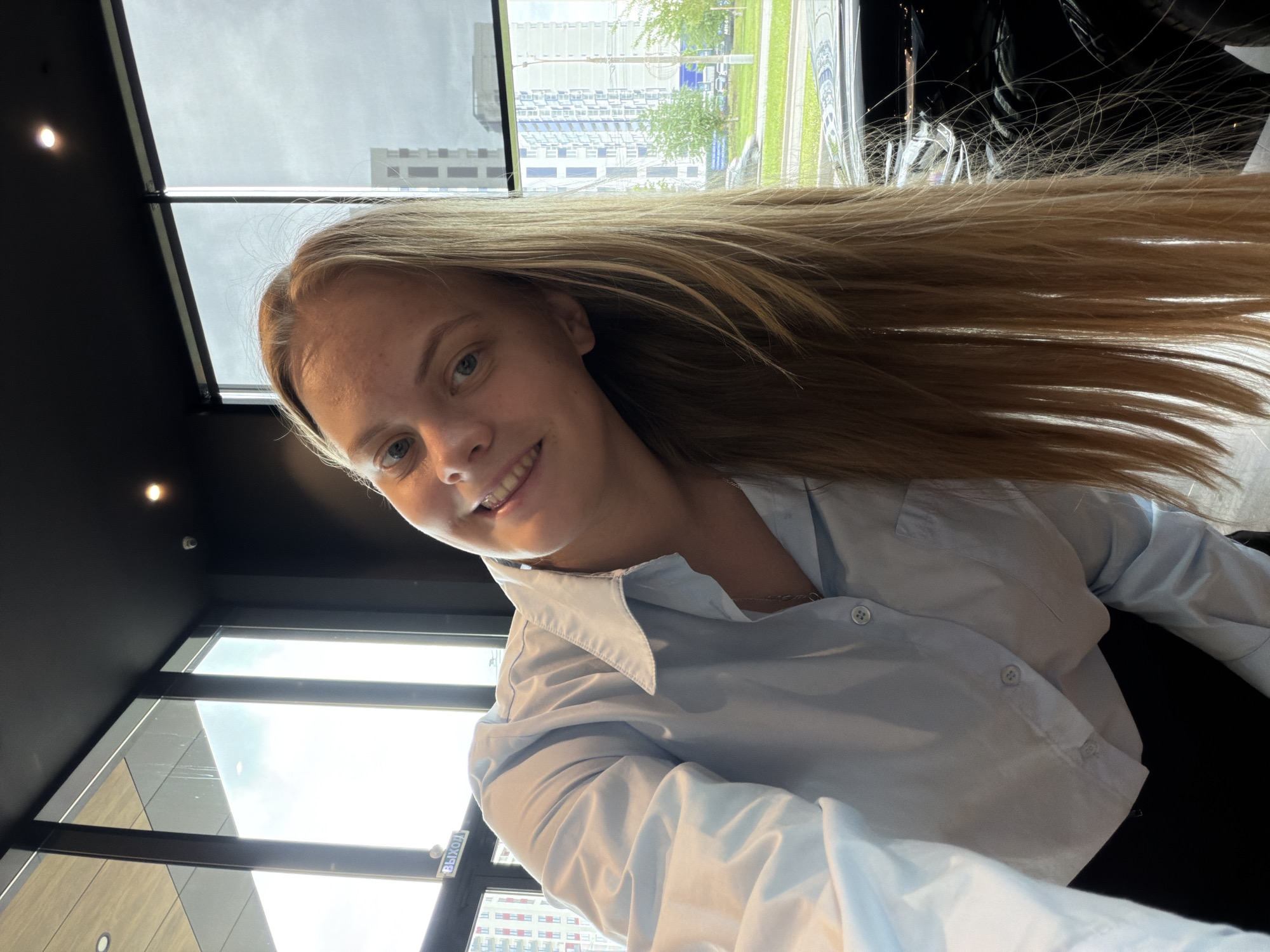 A woman with long blonde hair and wearing a light blue button-down shirt is indoors, possibly in an office or building in Izhevsk, Russia. She is smiling and looking towards the camera. The background includes a window showing buildings and greenery, typical of an urban environment. The interior has dark walls and possibly ceiling fixtures. The "выход" (exit) sign indicates she might be near an exit point.