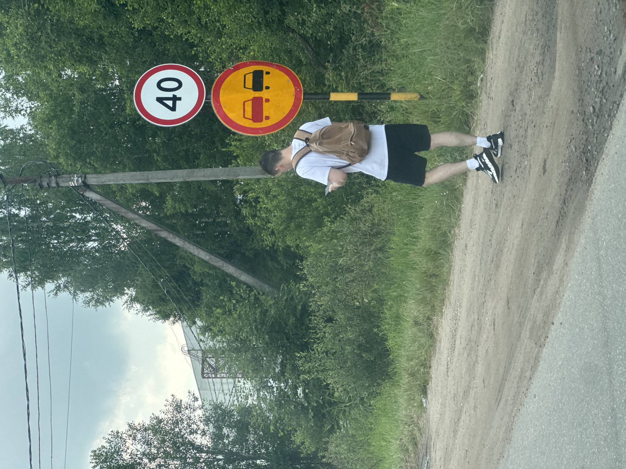 A young man wearing a white t-shirt, black shorts, white socks, and black and white sneakers stands on a dirt road. He has a brown backpack slung over his left shoulder and is looking down at his phone, which he holds in his right hand. The man is positioned to the right of two traffic signs. The first sign is circular with a red border and the number "40" in black in the center, indicating a speed limit of 40. The second sign is circular with a yellow background and a red border. It depicts two cars side-by-side with a red line through them, signifying "no overtaking." A black and yellow striped pole is visible behind these signs.
The scene is outdoors, with lush green trees and grass surrounding the dirt road. A power pole with wires stands to the left of the signs. In the background, a light-colored building structure with a metal framework is partially visible through the trees. The lighting suggests it is daytime, with a bright, overcast sky. The overall impression is a casual, everyday moment on a rural or suburban road in Novoe Devyatkino, Russia.