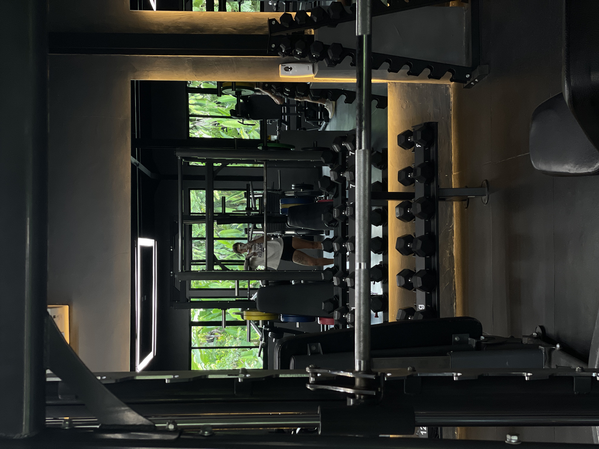 The image depicts the interior of a gym located in Mengwi, Indonesia. The gym features a dark, modern aesthetic with black equipment and walls, contrasted by warm, ambient lighting. In the foreground, a weight rack with dumbbells is visible. A woman, wearing a white t-shirt and black shorts, is reflected in a mirror, holding a barbell across her shoulders. She is standing in front of a squat rack, suggesting she is engaged in a weightlifting exercise. Behind her, large windows offer a view of lush green foliage, indicating the gym is situated amidst natural surroundings. The overall scene conveys a sense of focused activity within a well-equipped, contemporary fitness facility.