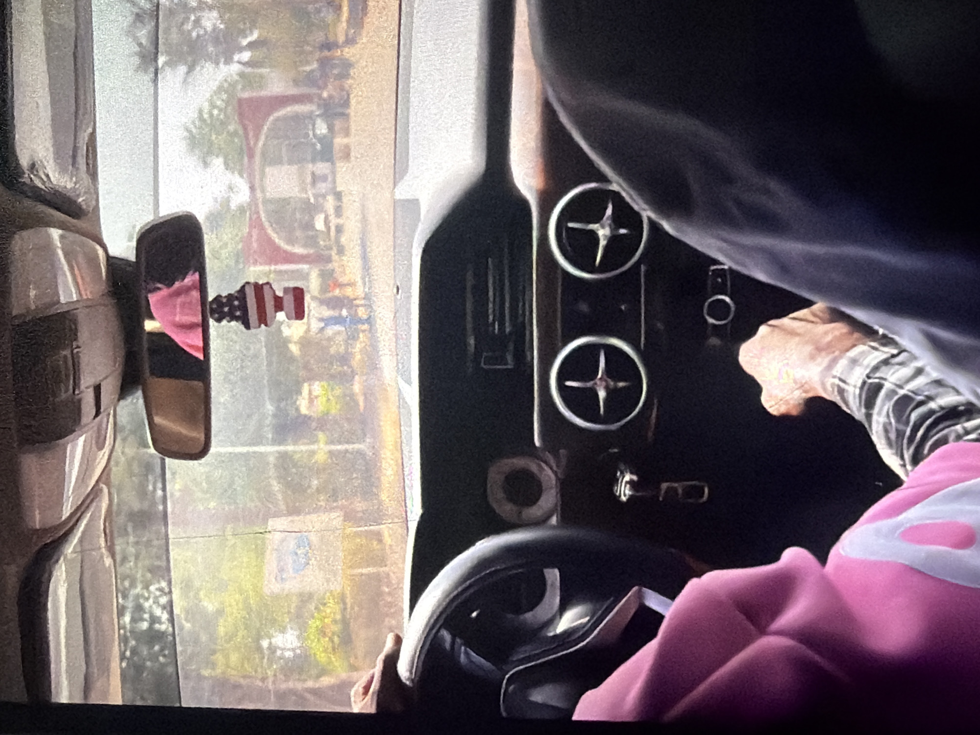 An image taken from inside a car shows the dashboard and the view through the windshield. A hand is visible on the steering wheel. A pink hoodie is worn by someone in the passenger seat. Outside, there is an archway and trees. A car air freshener in the shape of the American flag hangs from the rearview mirror. The scene appears to be a street in Bukuru, Nigeria.