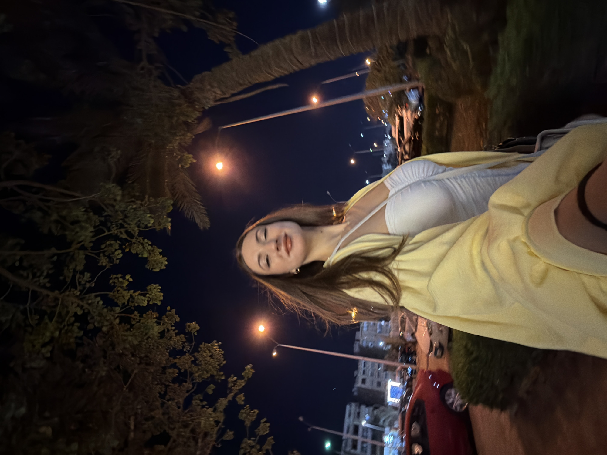 A young woman is taking a selfie at night on a street in the city of 6th of October, Egypt. She is wearing a white dress and a yellow cardigan. The background shows a street with palm trees, streetlights, and buildings. There are cars parked on the street. The woman is smiling and looking at the camera.