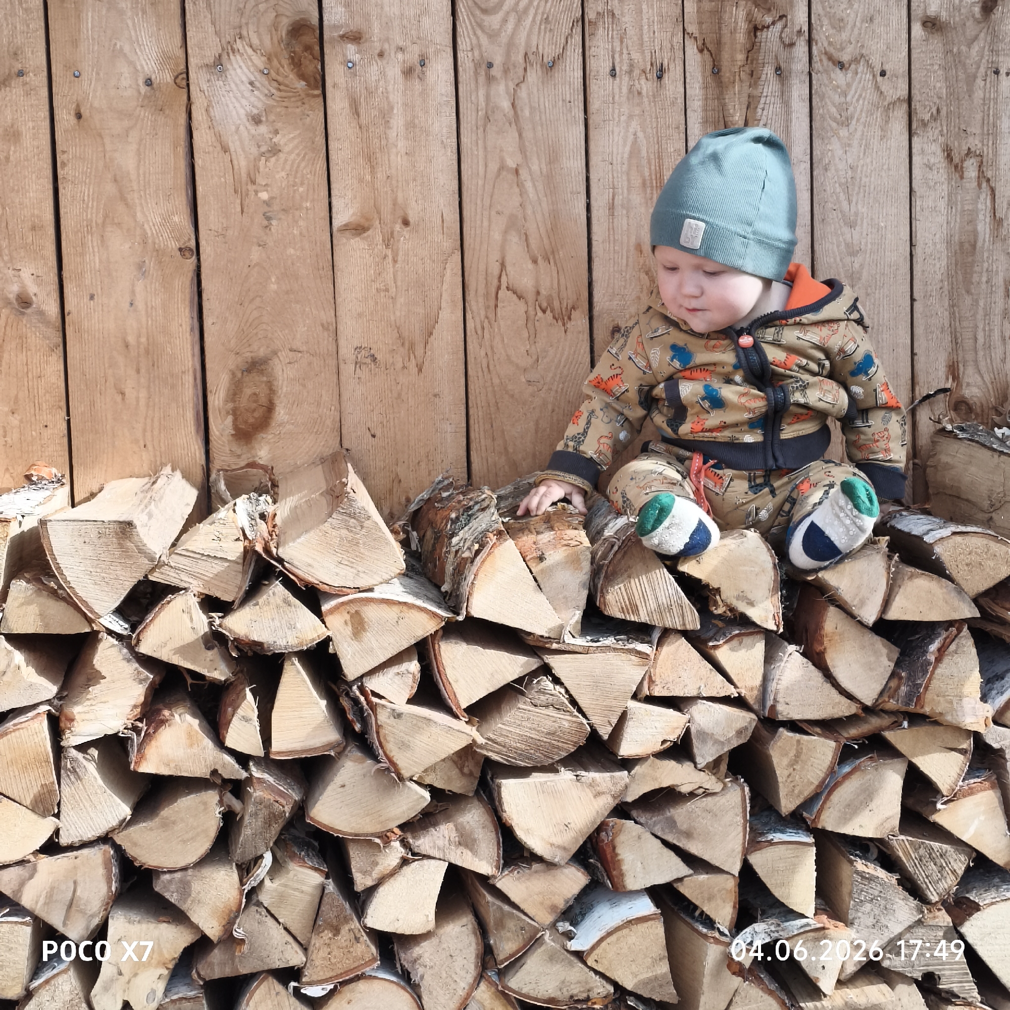 This outdoor photograph features a young child sitting atop a large stack of split firewood, positioned against a rustic wooden fence. The child, likely a toddler, wears a warm green knit beanie, a patterned full-body suit in brown and khaki tones with orange and blue accents, and colorful green, white, and blue booties. Their left hand gently rests on one of the birch logs as they gaze downwards.
The setting appears to be a backyard or a rural area in Chaikovsky, Russia. The background consists of a vertical plank wooden fence, showing visible knots and nails, suggesting unpainted or weathered wood. The firewood, primarily light-colored birch with peeling bark, is neatly stacked, creating a sturdy perch for the child.
The lighting in the image is bright and sunny, casting soft shadows, indicating a clear day. The time "17:49" in the timestamp confirms it was taken in the late afternoon. The child's warm attire suggests cooler temperatures despite the sunshine. The overall impression is one of peaceful observation or quiet play.
Visible text in the bottom left corner reads "POCO X7," indicating the camera device used. The bottom right corner displays the date and time: "04.06.2026 17:49."