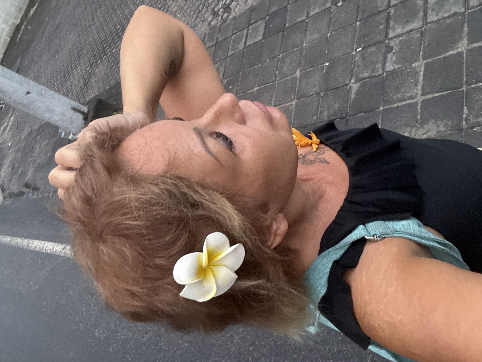 An overhead shot captures a woman relaxing outdoors, likely in Kuta, Indonesia. She is lying down or leaning back, gazing upwards and slightly to her right, with a serene and contemplative expression. Her light brown hair is adorned with a delicate white and yellow plumeria flower tucked behind her right ear.
She is wearing a black top with ruffled detailing around the neckline. A light blue or turquoise strap of another garment is visible near her left shoulder. On her left upper chest, a floral or tribal tattoo is visible beneath an orange, possibly floral, ornament or small necklace. Another faint tattoo can be seen on her right upper arm. Her right arm is bent, with her hand gently resting near her head.
The background shows dark grey cobblestone paving, suggesting a pedestrian area. Further away, a lighter grey surface, possibly asphalt or concrete, with a white line is visible, hinting at a road or parking space. A metal post or barrier is partially seen on the left side of the frame. The lighting appears soft, indicating either an overcast day or late afternoon/early evening. No text is visible in the image.
