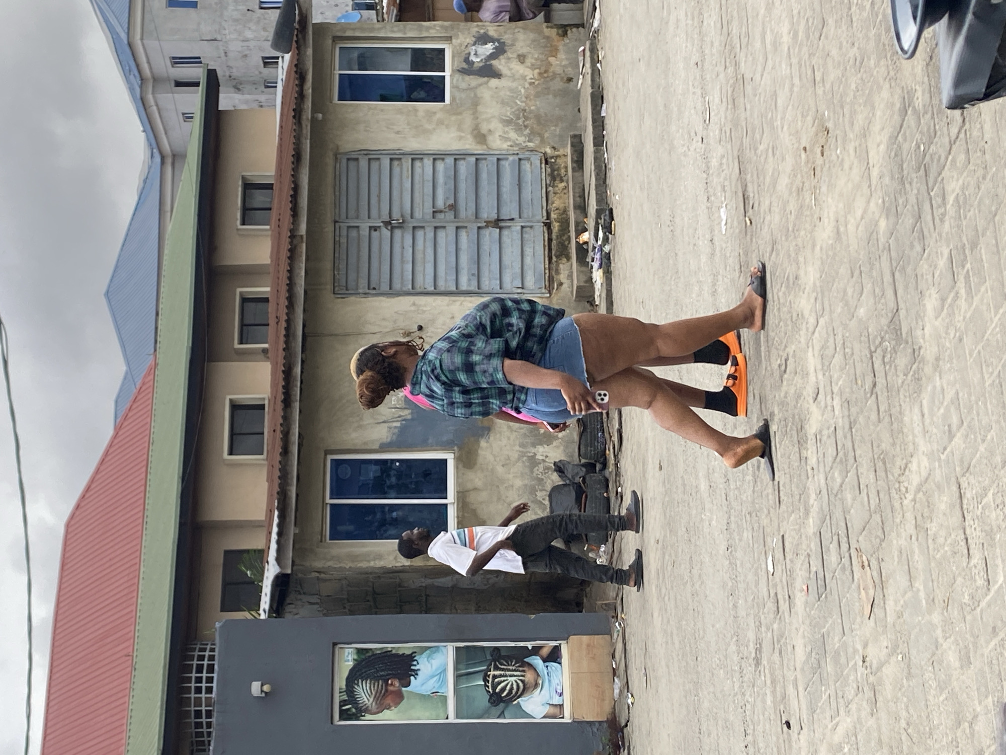 In an outdoor urban setting, two women are walking across a paved area. The woman in the foreground, on the right, is wearing a green and blue plaid short-sleeved shirt and denim shorts. She is walking away from the camera, and her right leg is bent. The second woman, behind and to the left of the first, is wearing a pink top and dark bottoms. A man in a white and orange striped short-sleeved shirt and dark pants is walking past a building. The building has a grey facade with two windows showing images of children with braided hair. Further down the street, another building has corrugated metal siding and blue-framed windows. In the background, there are multi-story buildings with various colored roofs, some of which appear to be under construction. The ground is a mix of interlocking pavers and concrete. Scattered debris is visible on the ground. The sky is overcast.