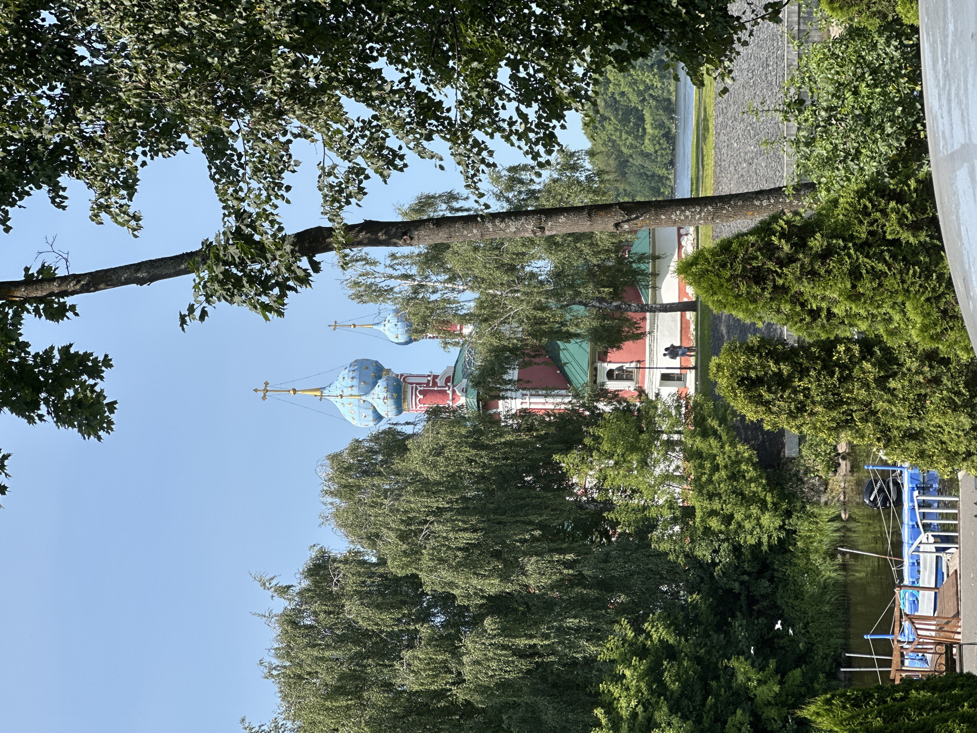 The image depicts a sunny day scene in Selivanovo, Russia, featuring a prominent red and white church with blue domes adorned with golden crosses, partially obscured by lush green trees. In the foreground, a body of water is visible with a blue and wooden dock. A small boat with an outboard motor is moored to the dock. Several birds are in flight above the water. The background shows a treeline and a body of water. Two individuals are standing near the church. The overall atmosphere is serene and picturesque.