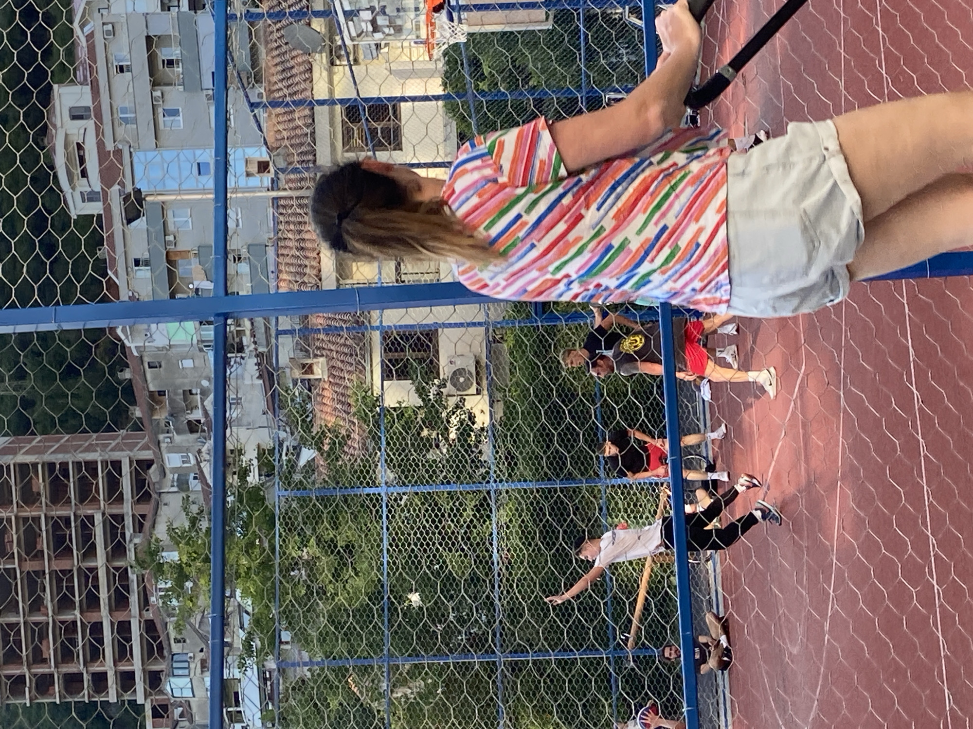 The image shows a fenced outdoor court in Budva, Montenegro. On the court, several people are playing some kind of field hockey or similar stick-and-ball game on a red surface marked with white lines. A woman in a multi-colored striped shirt and light-colored shorts stands close to the fence, partially obscuring the view. The surrounding environment includes buildings, trees, and a basketball hoop visible in the background. The photograph is taken from an elevated perspective.