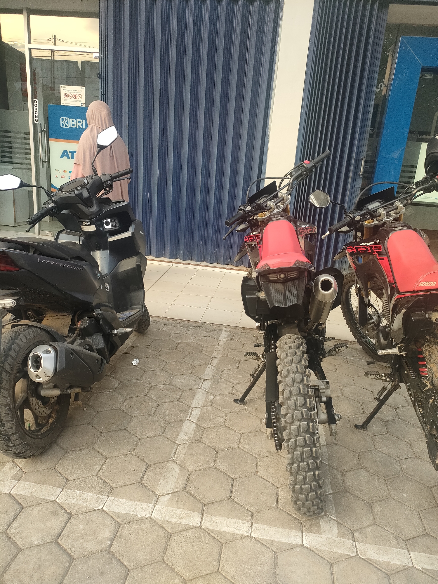 In front of a blue corrugated metal shuttered building, likely a bank branch in Sukoyoso, Indonesia, two motorcycles are parked on a hexagonal paver stone sidewalk. The motorcycle on the left is a black scooter with a silver exhaust. To the right, two red dirt bikes with black accents are parked. One dirt bike is closer to the camera and its rear tire, exhaust, and red seat are clearly visible. A person wearing a light pink hijab and a tan hijab is standing with their back to the camera, facing the building entrance, partially obscured by the black scooter. The building's entrance has a glass door and signage, including the "BRI" logo, indicating it is a Bank Rakyat Indonesia branch. The overall scene suggests a casual urban setting with vehicles parked outside a financial institution.