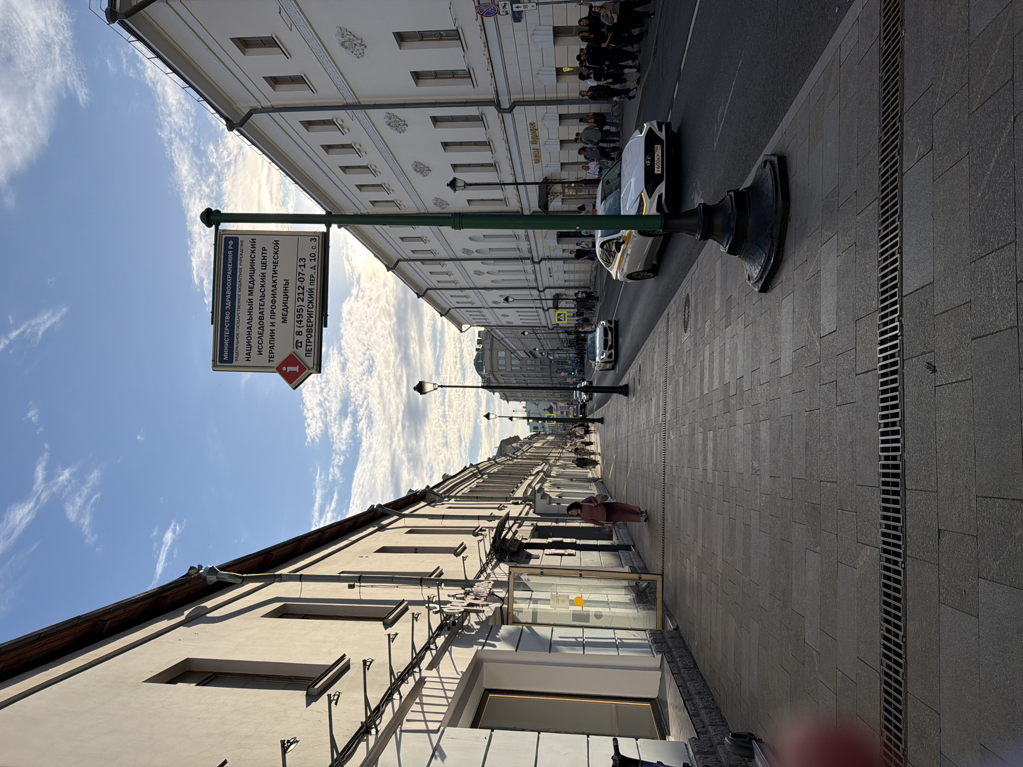 This image captures a street scene in Moscow, Russia. The view is oriented lengthwise along a narrow street lined with multi-story buildings. The architectural style suggests a historic urban area, with details like decorative moldings on the buildings.
On the right side of the image, pedestrians can be seen walking along the sidewalk. Parked cars are visible on the street, including a white car. A dark-colored curb drain runs along the edge of the sidewalk.
A sign is prominently placed on the left side of the image. The sign displays information in Russian, and references the Ministry of Health of the Russian Federation and the National Medical Research Center of Therapy and Preventative Medicine, located on Petrovsky Per., Building 10, Construction 3, phone number +7 495 212-07-13.
The sky is partly cloudy, indicating daytime. The image appears to have been taken at eye-level from a standing position.